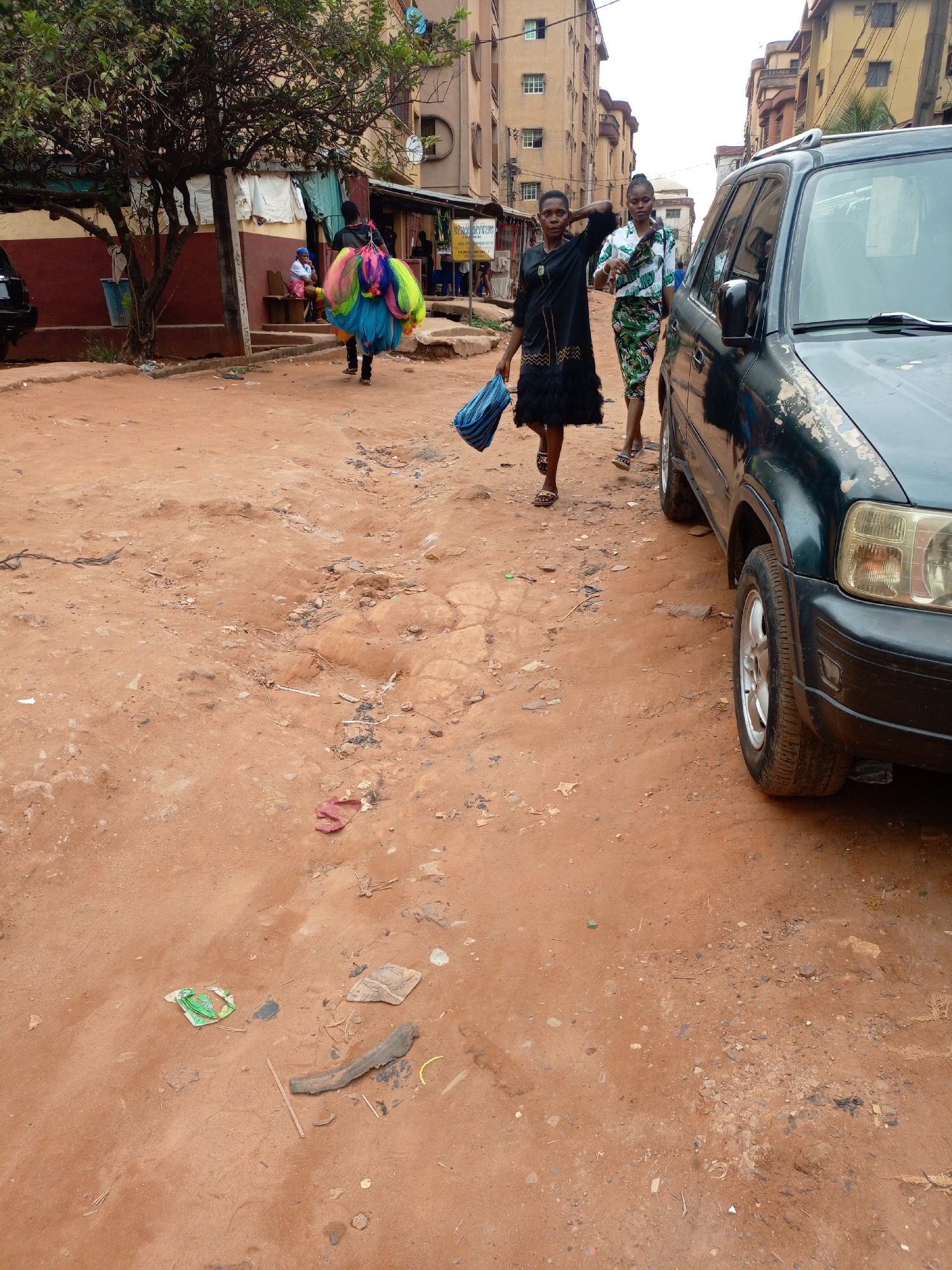 This media file captures a daytime street scene in Onitsha, Nigeria, featuring an unpaved, reddish-brown dirt road. The ground is uneven, marked with small potholes, and scattered with debris including plastic wrappers, small rocks, and pieces of wood.
In the foreground, a dark green SUV with chipped paint is parked on the right side of the road. Two women are walking towards the viewer on the dirt road; one wears a long black dress and carries a blue striped bag, while the other is dressed in a patterned white and green top and trousers. Further down the road, walking away from the viewer, an individual carries a large bundle of brightly colored mesh nets, likely a hawker. To the left, an elderly person is seated on a wooden bench beneath a large, leafy tree. Behind the tree, clothes are seen hanging out to dry against a building with a red lower wall.
The background is lined with several multi-story buildings, characterized by light-colored, faded facades, suggesting a residential or commercial urban setting. The sky is bright but overcast, indicating diffused daylight with no strong shadows. A partially visible sign in the mid-background appears to have the word "CO-OPERATIVE" visible. The scene depicts everyday activity on a bustling urban street.