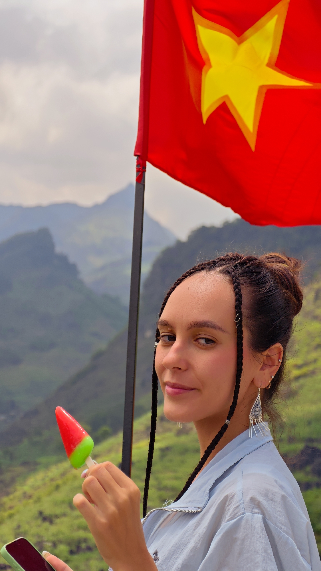 A woman stands in front of a red and yellow Vietnamese flag, holding a red and green popsicle and a cell phone. The background shows a mountainous landscape, consistent with the geography of Lũng Phìn Commune, Vietnam. The woman has dark hair styled in braids and a bun. She wears silver earrings and a light blue jacket.