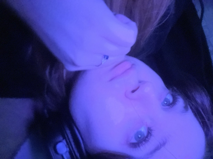 A young female is depicted in a close-up portrait, illuminated by a pervasive blue-purple light source. Her head is angled slightly to the right, with her blue eyes directed upwards and to the right. Her right hand is raised, with her chin resting on her knuckles. A silver-colored ring with a blue gemstone is present on her right ring finger. She wears dark over-ear headphones, with a light-colored ear cushion visible on the right side of her head. Her hair is dark brown or auburn. The background is dark and indistinct, suggesting an indoor environment. No specific location within Pushchino, Russia, is discernible from the visual information provided.