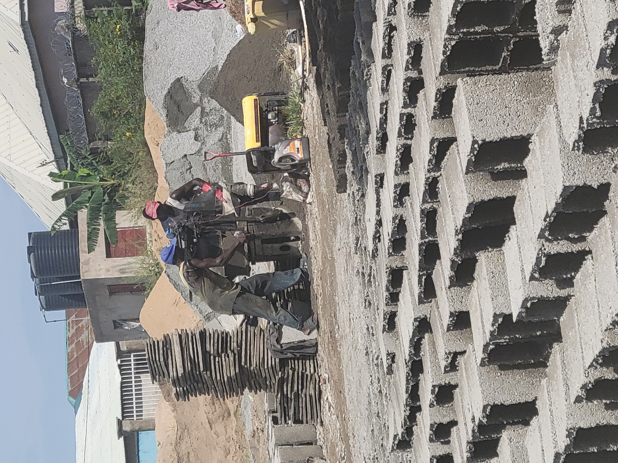 Two men are working at a construction site in Suleja, Nigeria, on a bright, sunny day. They are operating a block-making machine, which is positioned in the center of the frame. One man, wearing a blue cap, is standing to the left of the machine, facing it. The other man, wearing a red cap, is on the right, also facing the machine. Both men are dressed in work clothes.
The ground around the machine is covered with piles of sand and gravel, and there are stacks of what appear to be pre-made blocks. A yellow generator is situated behind the machine, and a red shovel leans against a pile of gravel nearby. In the background, buildings with corrugated iron roofs are visible, along with a water tank on top of one of the structures. Further back, there are trees and vegetation, and barbed wire fencing can be seen along a wall. The foreground is dominated by rows of freshly made concrete blocks, neatly arranged.