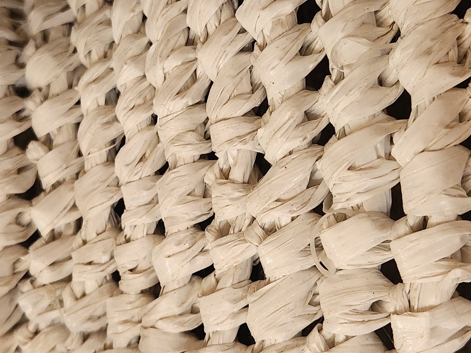 This is a close-up, angled shot of a woven material, likely a basket or a similar decorative item. The material appears to be made of thick, pale straw or raffia, tightly woven in a crisscross pattern. The lighting casts shadows that highlight the texture and depth of the weave, giving it a tactile quality. Some strands of the material show slight variations in color and texture, with minor specks of what could be dirt or natural imperfections in the straw. The overall impression is one of rustic craftsmanship. No people, activity, text, or specific location cues are visible beyond the general context of the United States of America, Maitland. The time of day and weather are not discernible from the image.