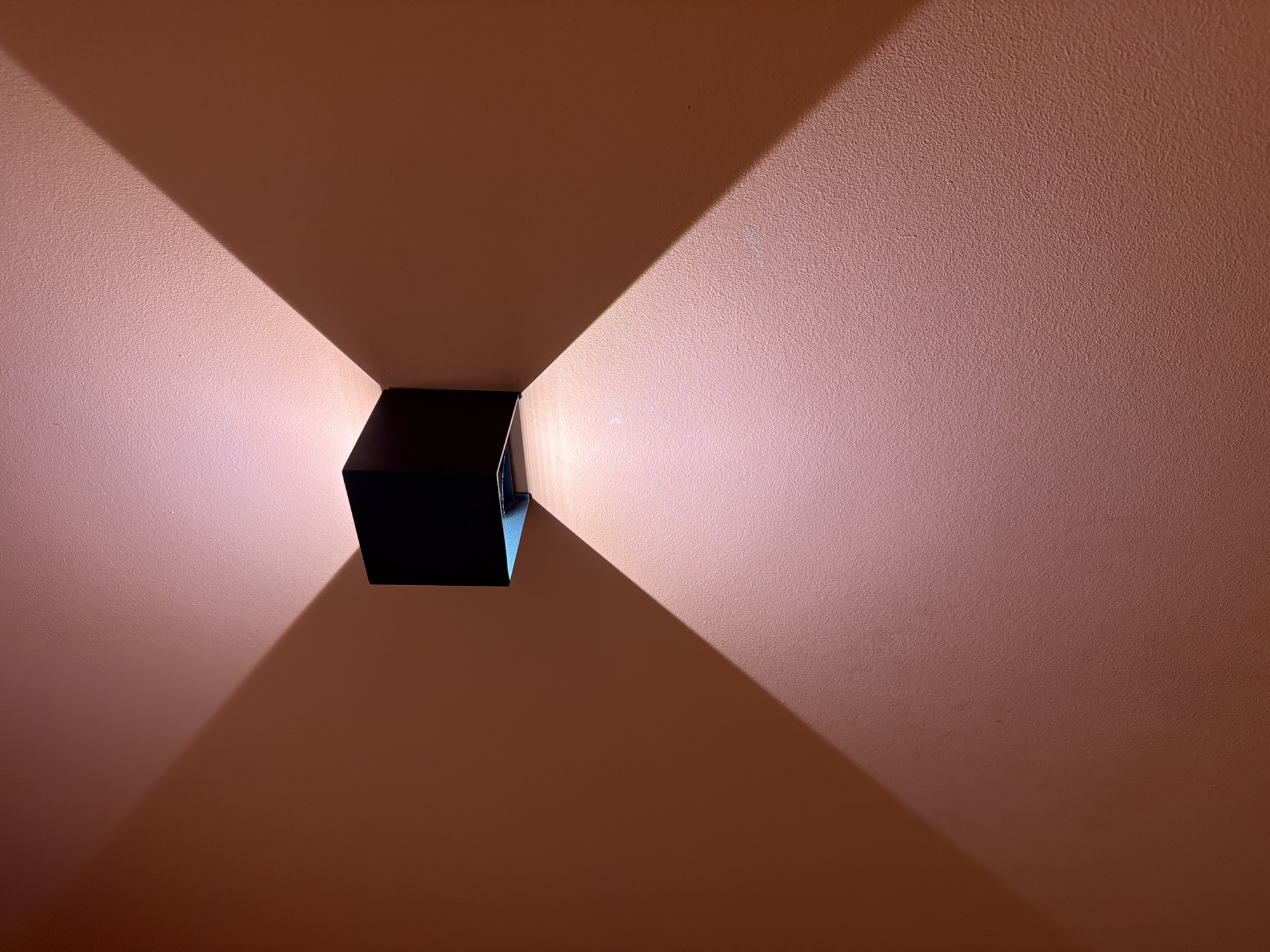 The image shows a black, rectangular wall sconce mounted on a peach-colored wall. The sconce emits light upwards and downwards, creating two triangular light patterns on the wall. The wall has a textured surface. The location is unknown.