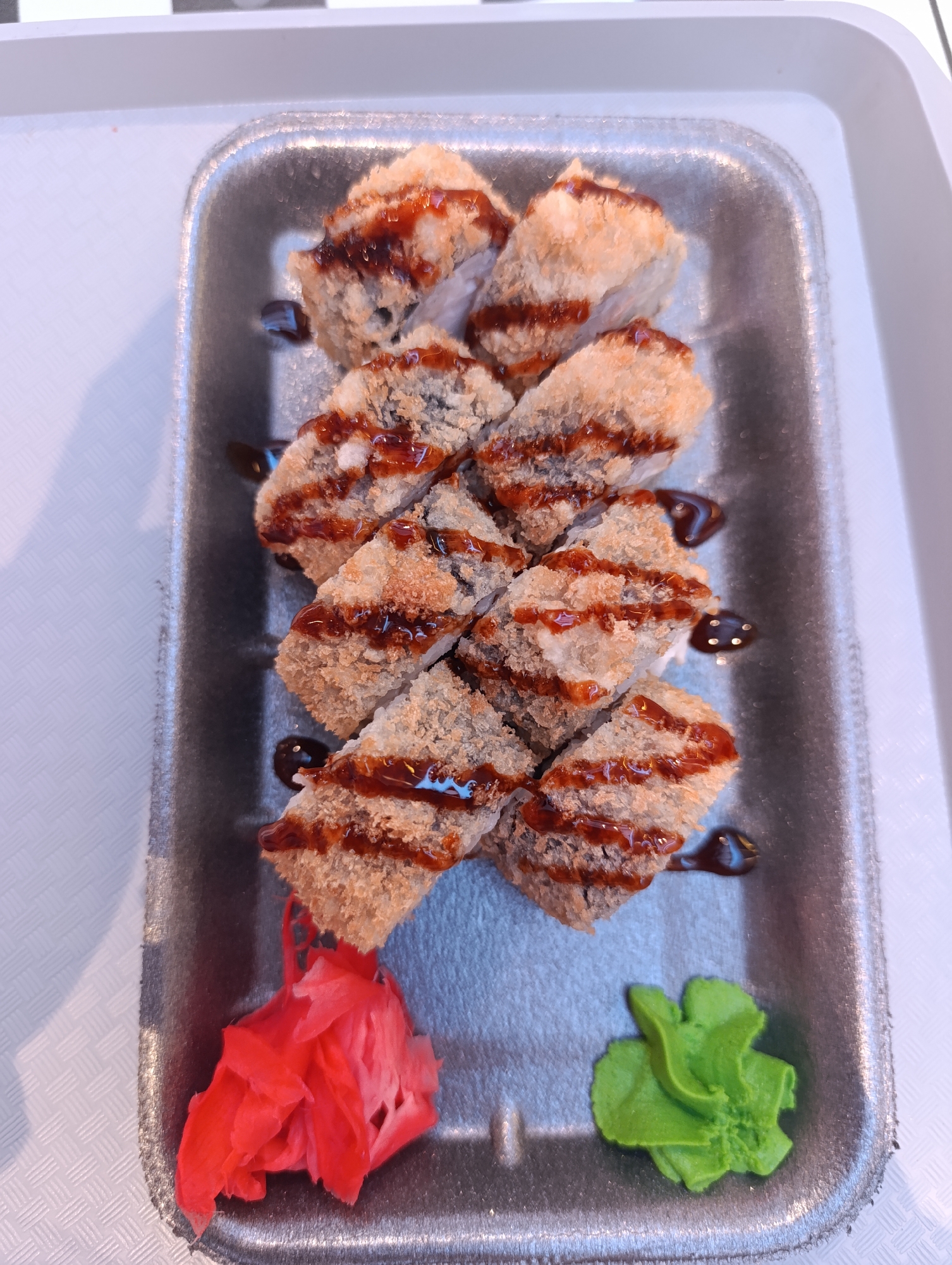The image depicts a takeaway container of sushi, arranged on a white table. The sushi rolls appear to be fried and are drizzled with a dark sauce. The container also holds a portion of pickled ginger and wasabi. The image was taken in Moscow, Russia.