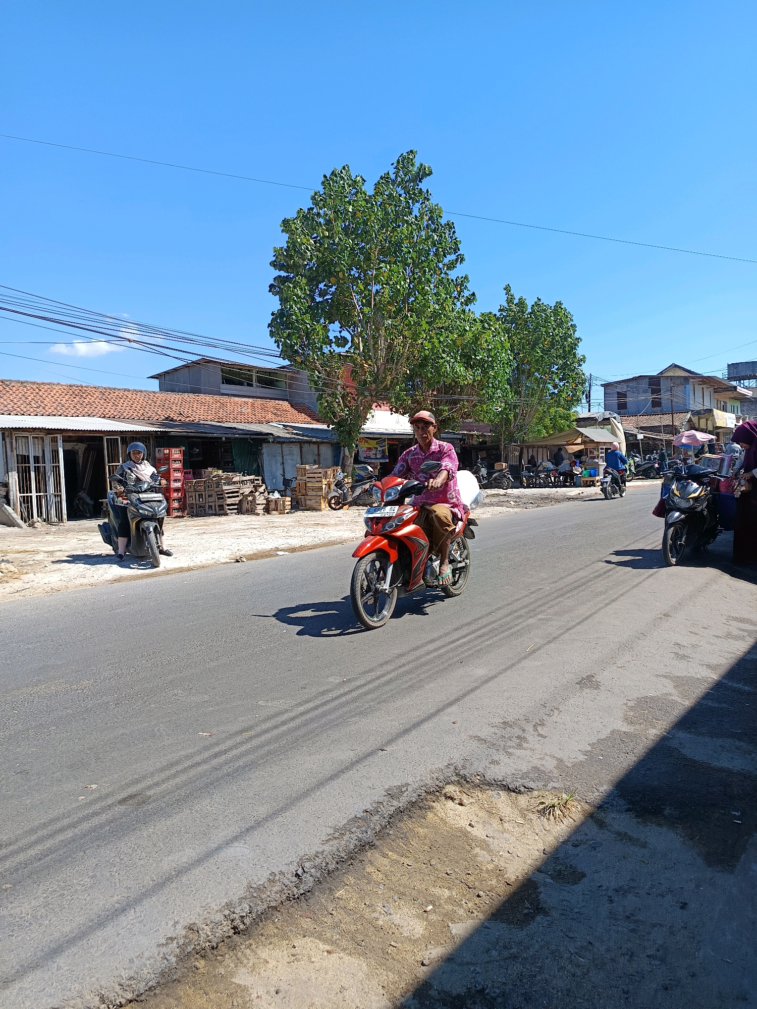 The image depicts a street scene in Blega, Indonesia, under a clear blue sky. The foreground shows an asphalt road with visible tire marks and shadows cast by overhead wires. On the left side of the road, a woman wearing a hijab and a black and white patterned shirt rides a black motorcycle. Behind her, a building with a tiled roof and a metal awning appears to be a shop or market, with wooden pallets and crates stacked outside.
Further down the road, a man wearing a red and white patterned shirt and a red cap rides an orange motorcycle. To the right of the road, a black motorcycle is parked, and a woman in a dark purple hijab and a dark top stands near it, possibly engaged in a transaction. In the background, other buildings and trees are visible, suggesting a semi-urban or rural environment. The overall scene conveys a typical day in an Indonesian town.