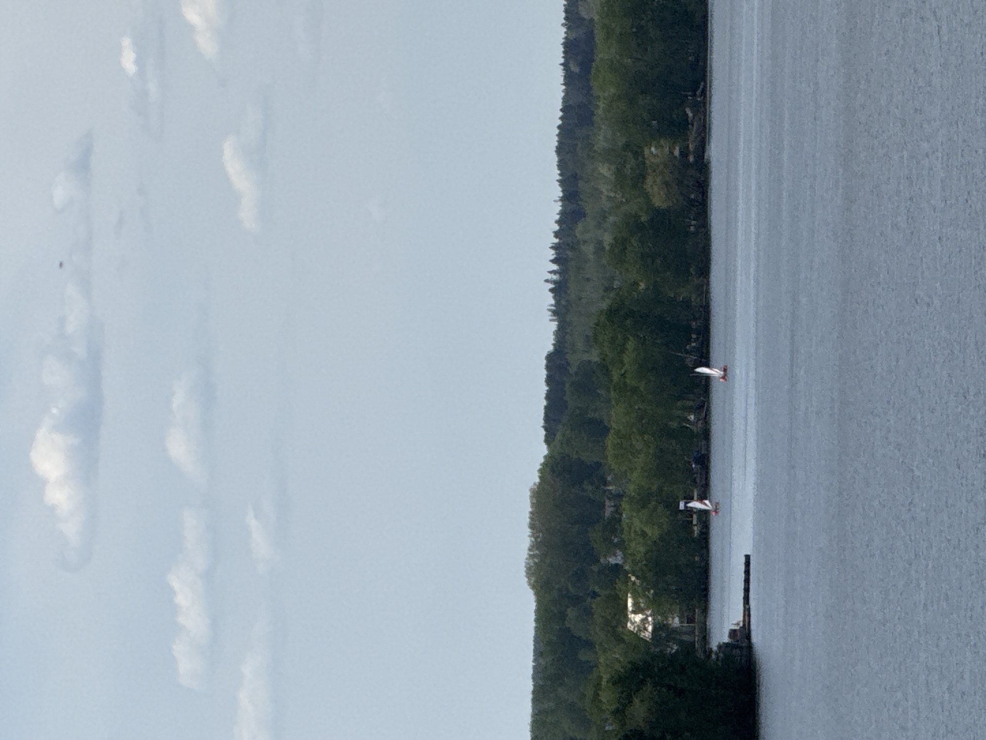 The image shows a body of water, possibly a lake or wide river, adjacent to a shoreline with dense trees. The sky has scattered clouds. Two boats with white sails are visible on the water, along with what may be a dock extending into the water. The image is taken during daylight. There is not enough information in this image to determine if the location is Votkinsk, Russia.