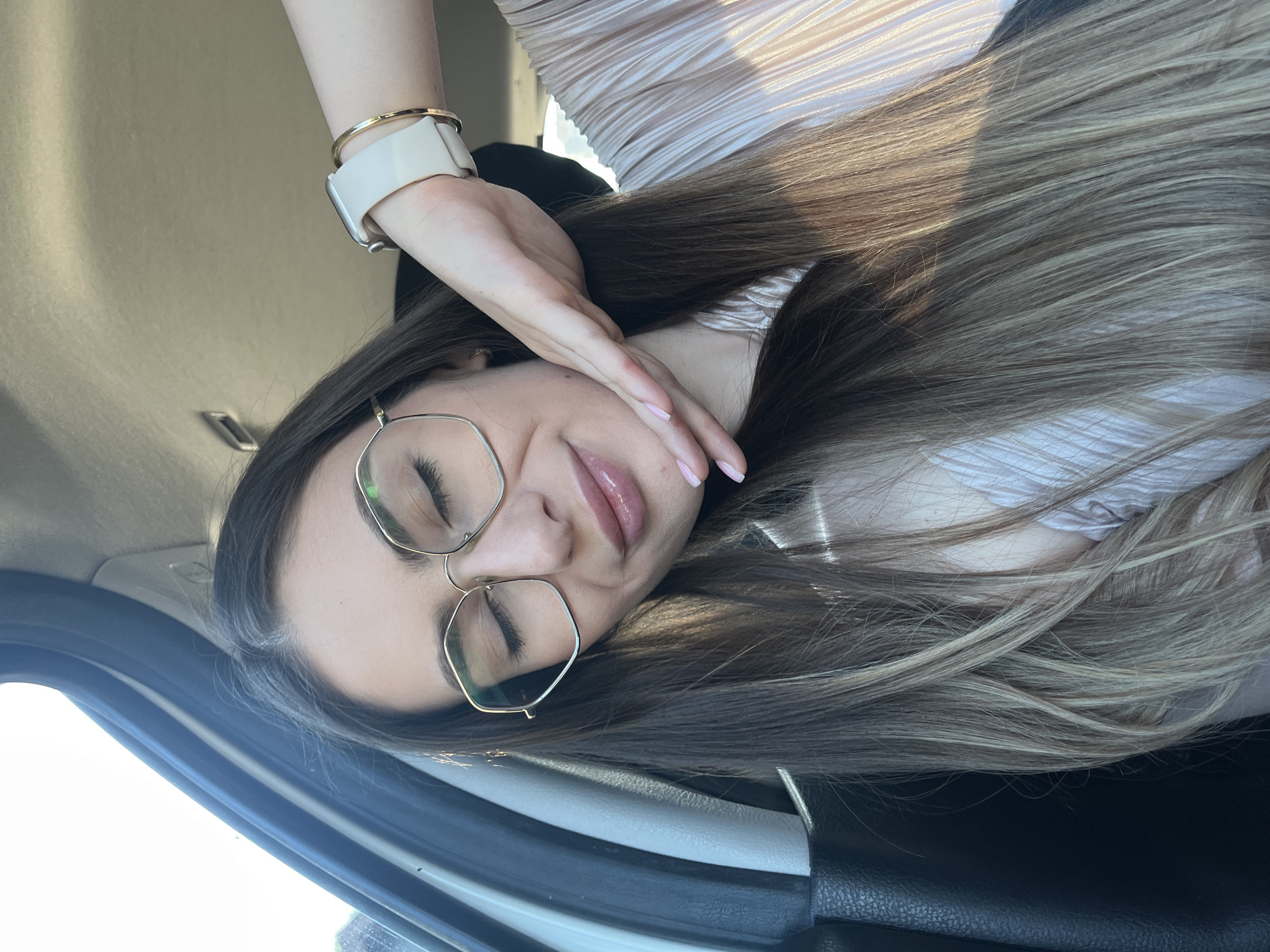 A woman with long, highlighted hair and gold-rimmed glasses is sitting in a car in Unknown, Unknown. She is wearing a light-colored, pleated top and a white smartwatch with a gold bracelet. Her hand is resting near her face. The car's interior is visible, including the door and seat.