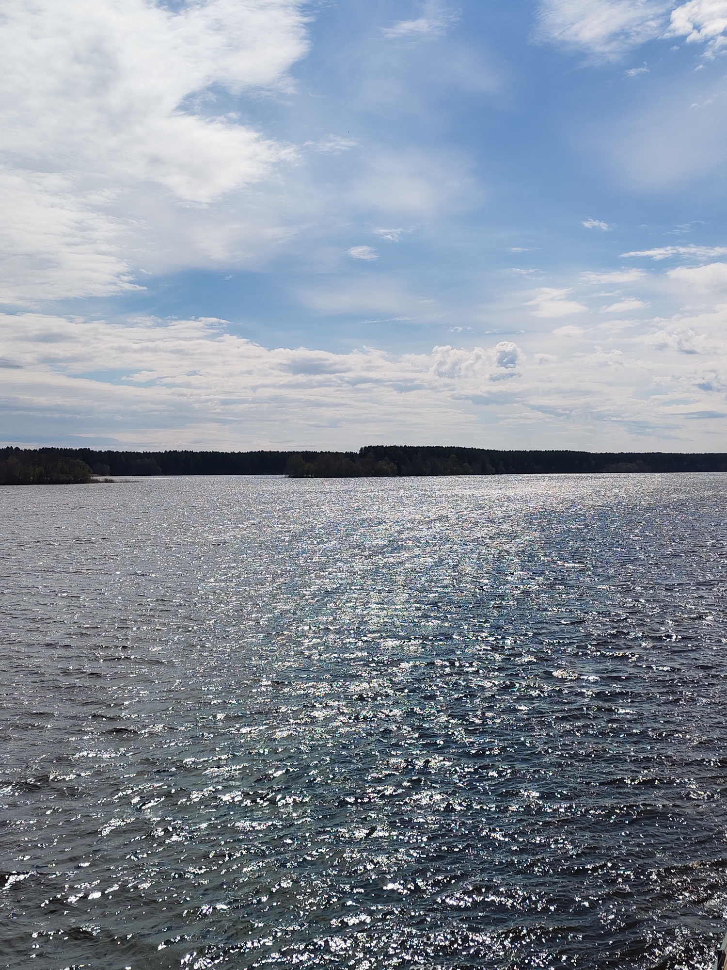 The image shows a body of water with a dark shoreline in the distance. The sky is blue with scattered white clouds. The water surface is textured with visible ripples and a sparkling effect, likely due to sunlight reflecting off the waves. The location is unspecified.