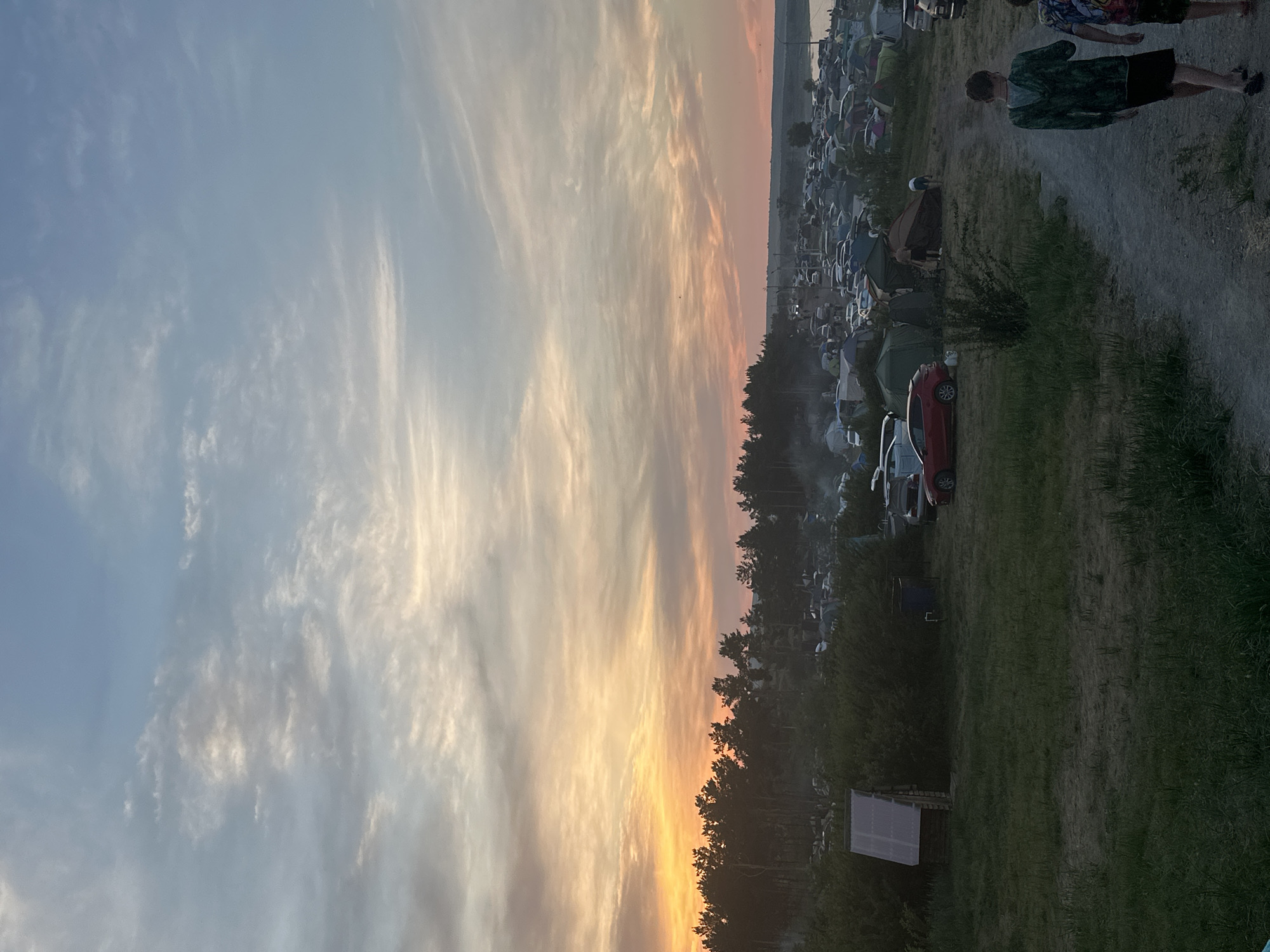 The image depicts a large outdoor gathering, possibly a festival or camping event, at dusk in the vicinity of Жемчужный, Russia. In the foreground, two young males, dressed casually, walk away from the camera on a dirt path. The middle ground features a campsite with numerous tents and cars, situated behind a line of trees. The sky above is filled with dramatic clouds, illuminated by the setting sun, casting orange and pink hues. The overall scene suggests a lively event taking place in a natural setting.