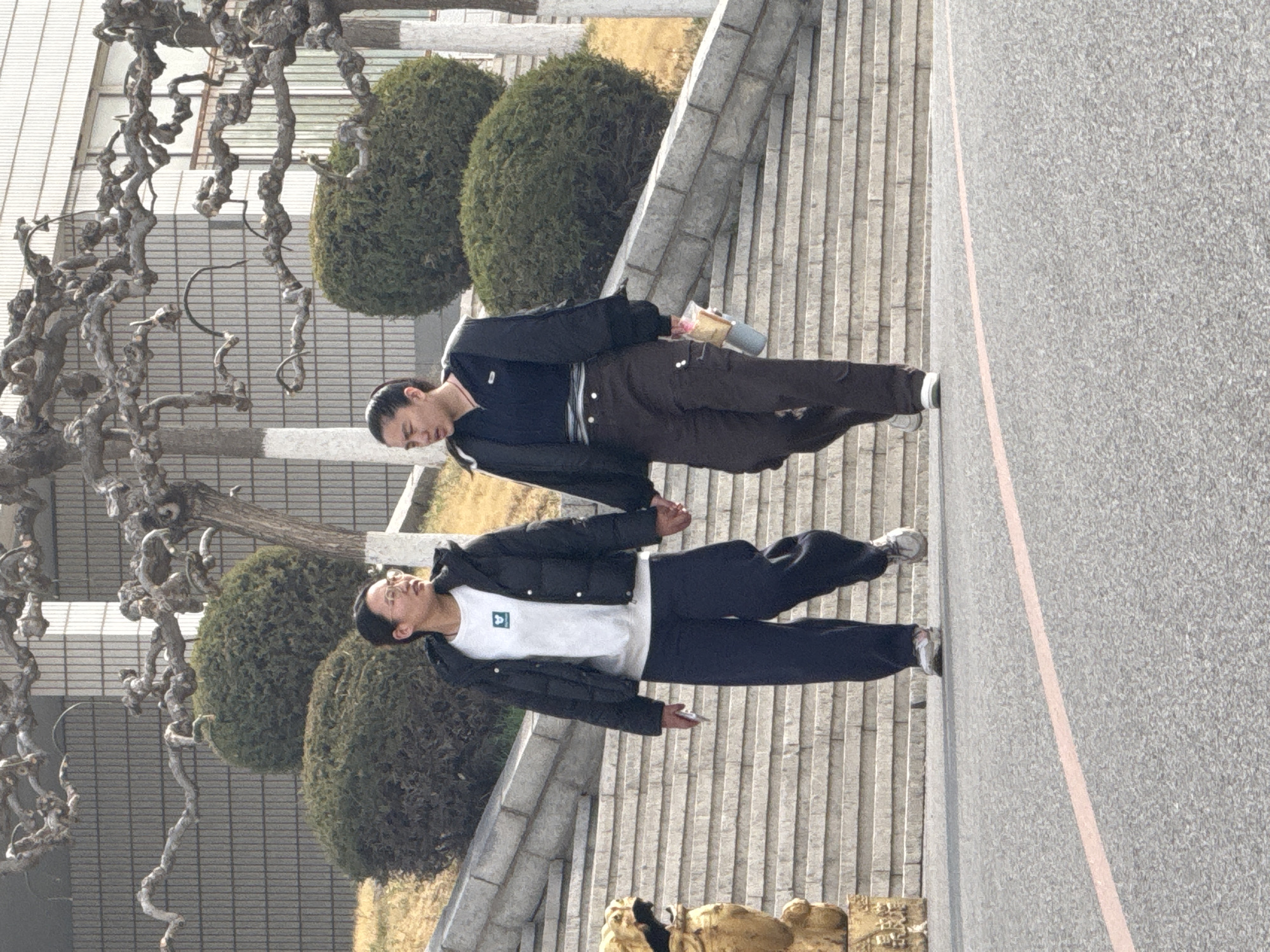 Two East Asian men walk side-by-side on a paved area with adjacent stone steps in Zhongtun, China. One wears a dark jacket over a white shirt and dark pants; the other wears a dark jacket over a dark shirt and brown pants and holds a cylindrical object. Behind them is a row of rounded bushes and a large, gnarled tree. The background includes a light-colored building. A golden statue is visible at the bottom of the image. A reddish-pink line runs along the right edge of the pavement.