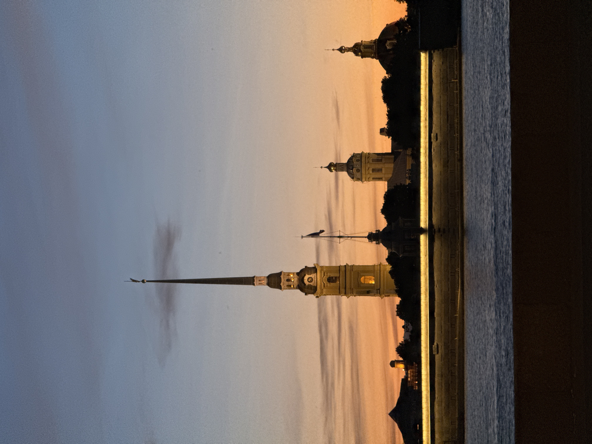 The image captures a sunset over the Peter and Paul Fortress in Saint Petersburg, Russia. The sky is a gradient of blues and oranges, with wispy clouds. The most prominent structure is the tall, slender spire of the Peter and Paul Cathedral, topped with a golden angel. To the right of the cathedral, a smaller spire with a flag is visible. Further to the right, two domed buildings, likely part of the fortress complex, are silhouetted against the sky. In the foreground, a wide river flows, reflecting the colors of the sunset. A stone embankment with illuminated lighting runs along the water's edge. The scene is serene and atmospheric, showcasing the historical architecture of the fortress against a natural backdrop.