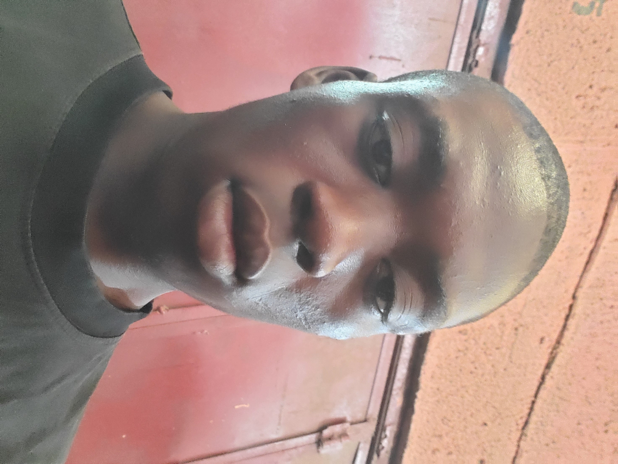 A medium-shot portrait of a dark-skinned man is shown against a reddish-orange, textured background. The man has short, dark hair and is wearing a black crew-neck shirt. His gaze is directed slightly downward. The photo appears to have been taken indoors with lighting that causes a sheen on the man's skin. The location is Unknown, Unknown.