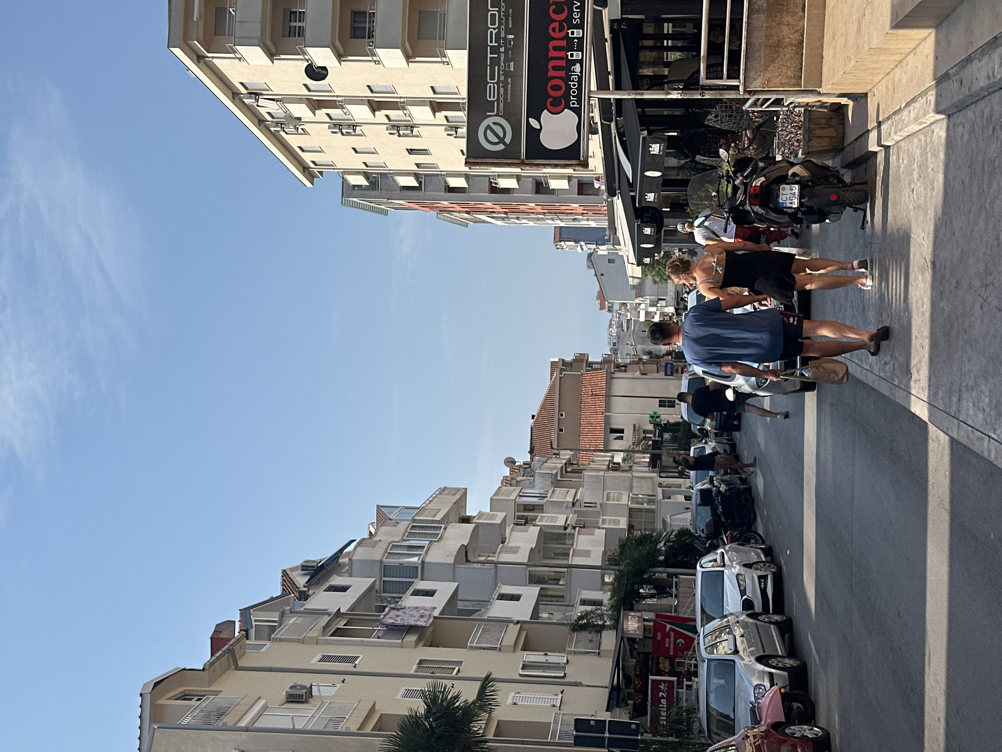 The image depicts a street scene in Budva, Montenegro, on a clear, sunny day. The viewpoint is from a low angle, looking down a moderately wide street lined with multi-story buildings on both sides. The architecture suggests a European coastal town with apartments or commercial spaces on the ground floor.
On the left side of the street, several apartment buildings with balconies, air conditioning units, and shuttered windows are visible. A palm tree is present on the far left. Cars are parked along the curb, including a silver station wagon, a silver hatchback, and a red vehicle. A shop sign reads "Stella 2".
On the right side, more apartment buildings are visible, with a prominent sign for "LECTRON GADGET STORE & IT SOLUTIONS" and "connect". Below these signs are awnings for businesses. A scooter with license plate "CT AG46" is parked on the sidewalk.
Several people are walking on the sidewalk. In the foreground, a couple is walking away from the camera, the man in a blue t-shirt and black shorts, and the woman in a black dress. Another person in a white shirt and red shorts is walking further down the street. Other individuals are partially visible in the background.
The street is paved with asphalt, and the sidewalk is made of concrete. The sky is a clear, bright blue with a few wispy clouds. The overall impression is a vibrant, sunlit urban environment in a Mediterranean setting.