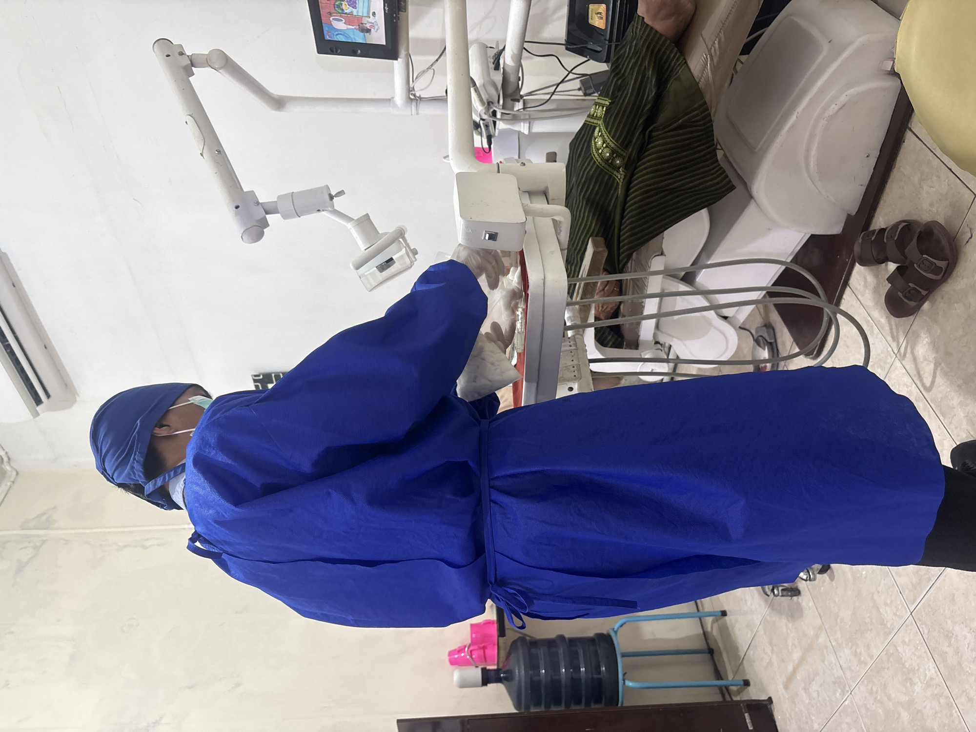 The image portrays an indoor scene within a medical or dental clinic, located in Suko, Indonesia. The central figure is a medical professional, viewed from behind, wearing a full protective ensemble: a blue disposable gown, a blue scrub cap, a white face mask, and white gloves. They are positioned in front of and interacting with white medical equipment, which includes an articulated arm typical of an examination light or tool delivery system in a dental setup.
In the background, a patient is partially visible, seated or reclined in a medical chair. This individual wears a dark green garment with a textured pattern, possibly traditional clothing. Dark sandals are visible on the light-tiled floor near the patient's feet. The clinic walls are white, with a mounted black flat-screen monitor showing a cartoon or graphic in the upper right. Exposed electrical wires are present near the monitor. Other objects include a large clear plastic water bottle on a blue metal stand and a small pink container on the floor. An air conditioning vent is partially visible on the left wall.
The activity depicted is likely the preparation for or execution of a medical or dental procedure, with the professional attending to the equipment. The setting suggests a local community clinic environment. No discernible text is visible in the image.