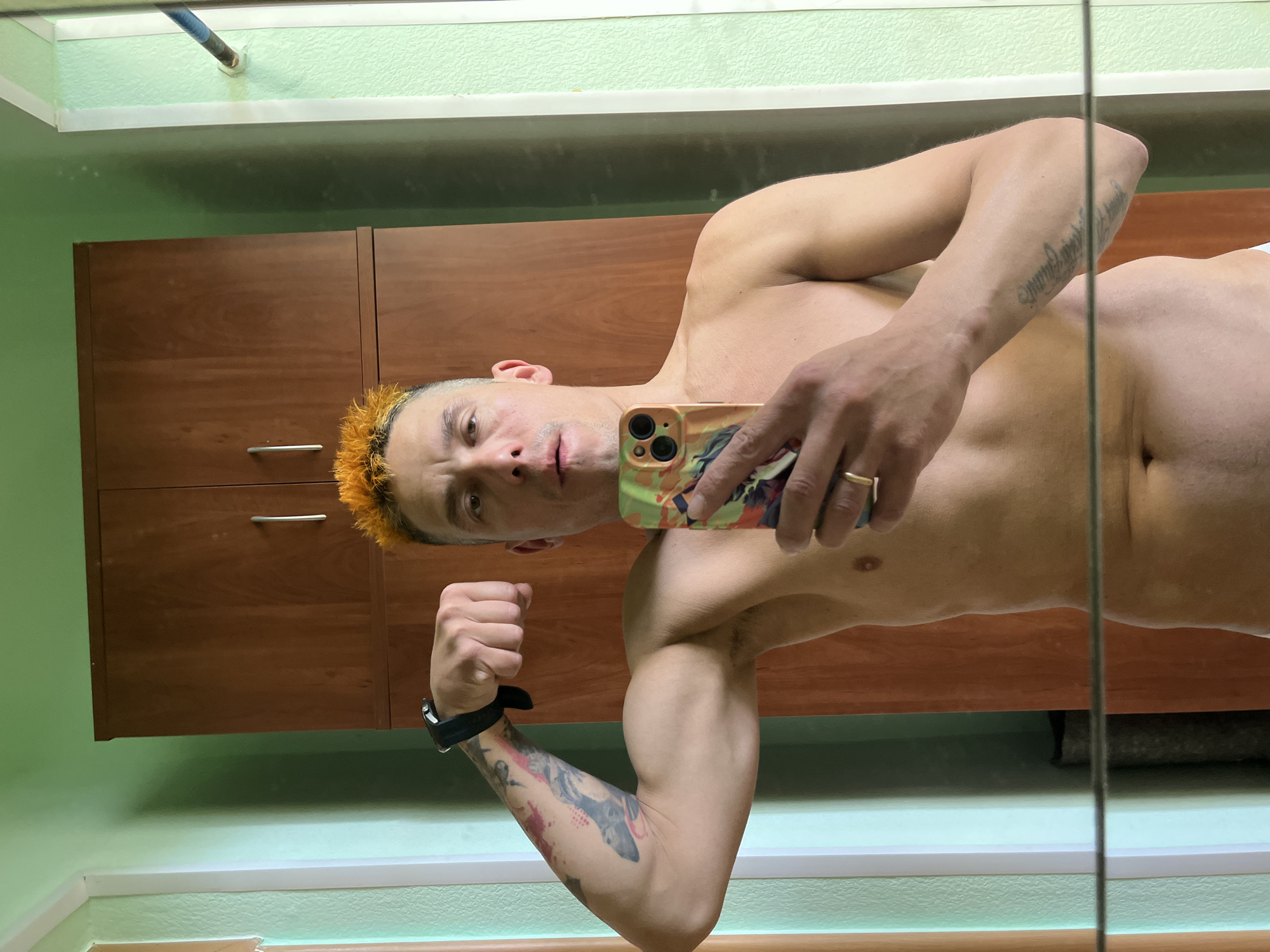 A bare-chested man is taking a selfie in a bathroom or changing room. He is facing the mirror, holding a smartphone in his right hand. He has short, orange hair and tattoos on both arms. He is flexing his left arm. Behind him are brown cabinets and light green walls. The location is not evident from the image.