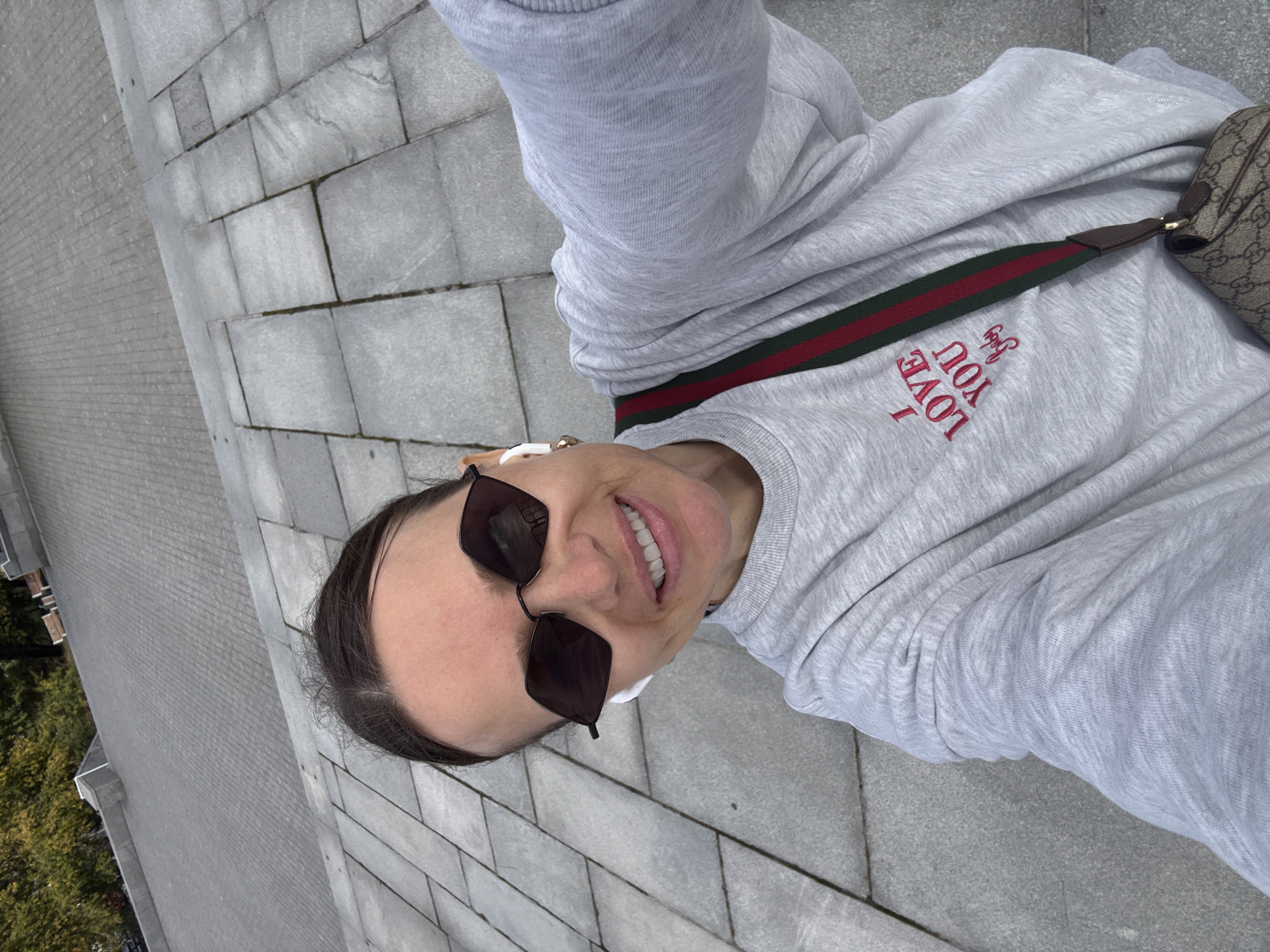 In a high-angle selfie, a woman with dark hair pulled back, wearing sunglasses and white earbuds, smiles broadly while standing outdoors on a paved walkway. She is dressed in a gray sweatshirt with red text that reads "I LOVE YOU Baby" and is carrying a brown patterned crossbody bag with a red and green striped strap. The background consists of gray stone pavement and some foliage. This scene was captured on a paved area near a park in Moscow, Russia, with the soft sunlight suggesting it is daytime.