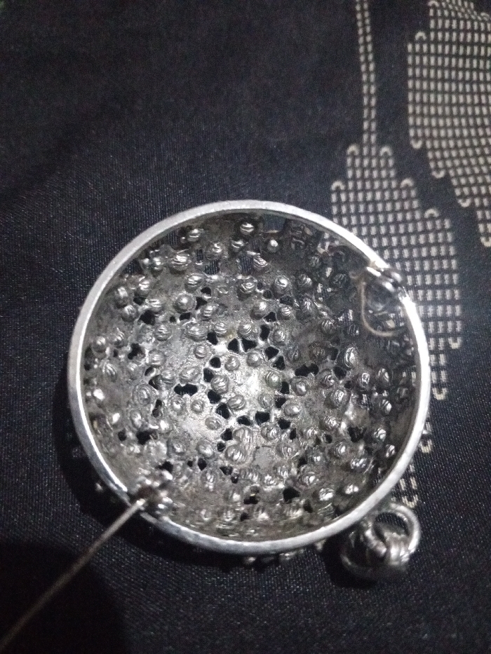 A single, silver-toned, hemispherical metal object, approximately 6 cm in diameter, is depicted against a dark, patterned fabric surface. The object features an intricately perforated design with a textured surface composed of numerous raised, granular elements. A thin metal wire extends from the object's lower-left periphery, and a small metal jump ring is attached to its upper-right side. The background fabric is dark black or navy with a repeating lighter-colored, stylized pattern. The scene is located in Hasanpur, India. No individuals are present.