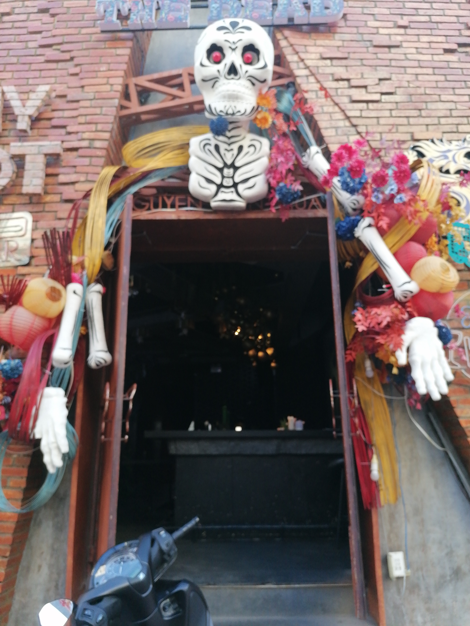 A decorated doorway is the focal point of the image. The doorway is adorned with a large, white skeleton figure with red eyes and colorful floral arrangements. To the left of the doorway is a sign with the words "MY ST" visible. The building appears to be constructed of red brick. A black scooter is parked at the entrance. Inside the doorway, a bar area with dim lighting can be seen. Based on the sign "SUYEN" above the doorway, it is likely that this is a restaurant or bar. The location is in Nha Trang Ward, Vietnam.