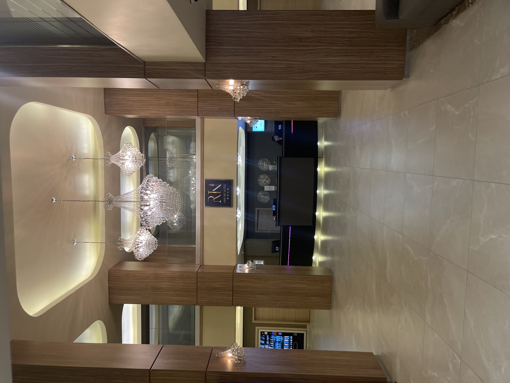 This is an interior shot of the reception area of the RIN Airport Hotel in Otopeni, Romania. The viewpoint is taken from the entrance, with a long corridor leading to the front desk. The reception desk is dark and staffed by an individual. Above the desk are multiple clocks displaying different time zones. The flooring consists of light-colored tiles. Elaborate crystal chandeliers hang from the ceiling. The walls and support structures are clad in wood veneer panels. A sign with the hotel's name and logo is visible behind the chandeliers. A refrigerated display case with beverages is positioned near the bottom of the frame.