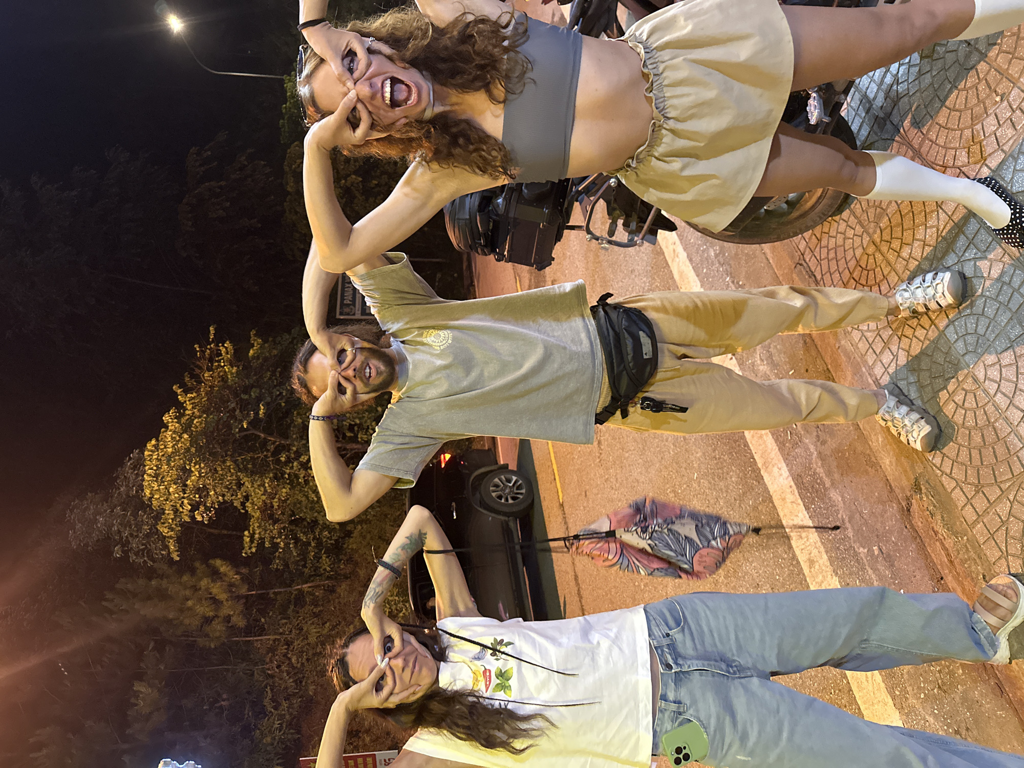 The image displays three young adults captured in a playful moment outdoors at night, likely in Mù Cang Chải, Vietnam. The scene is illuminated by streetlights, with dark trees and a road visible in the background, along with a tiled sidewalk in the foreground.
The topmost person, a woman with long, curly brown hair, is seated on a black motorbike. She wears a gray crop top and a light beige ruffled skirt, accessorized with white socks and black-and-white polka-dotted shoes. She makes an exaggerated, humorous face, stretching her eyes wide with her fingers and opening her mouth in a broad, playful expression.
Below her is a man with a beard, dressed in a light gray t-shirt with a graphic (partially legible, possibly "BEER") and light brown cargo pants, paired with white sneakers. He wears a black fanny pack across his waist. Mimicking the woman's pose, he also stretches his eyes with his fingers and smiles broadly. His arm appears to be around the waist of the woman above him.
The third person, another woman with long dark hair, is positioned at the bottom. She wears a white t-shirt adorned with colorful vegetable graphics (such as chili peppers and herbs) and light blue jeans, complemented by light-colored sandals. A green-cased smartphone is visible in her back pocket. She, too, is making a playful face by stretching her eyes with her fingers.
Notable details include a parked black car and a partially visible patterned umbrella leaning against something on the sidewalk. A faint white sign with "PANAY V" can be seen behind the man. The overall atmosphere of the image is joyful and informal, highlighting a fun interaction among the group.