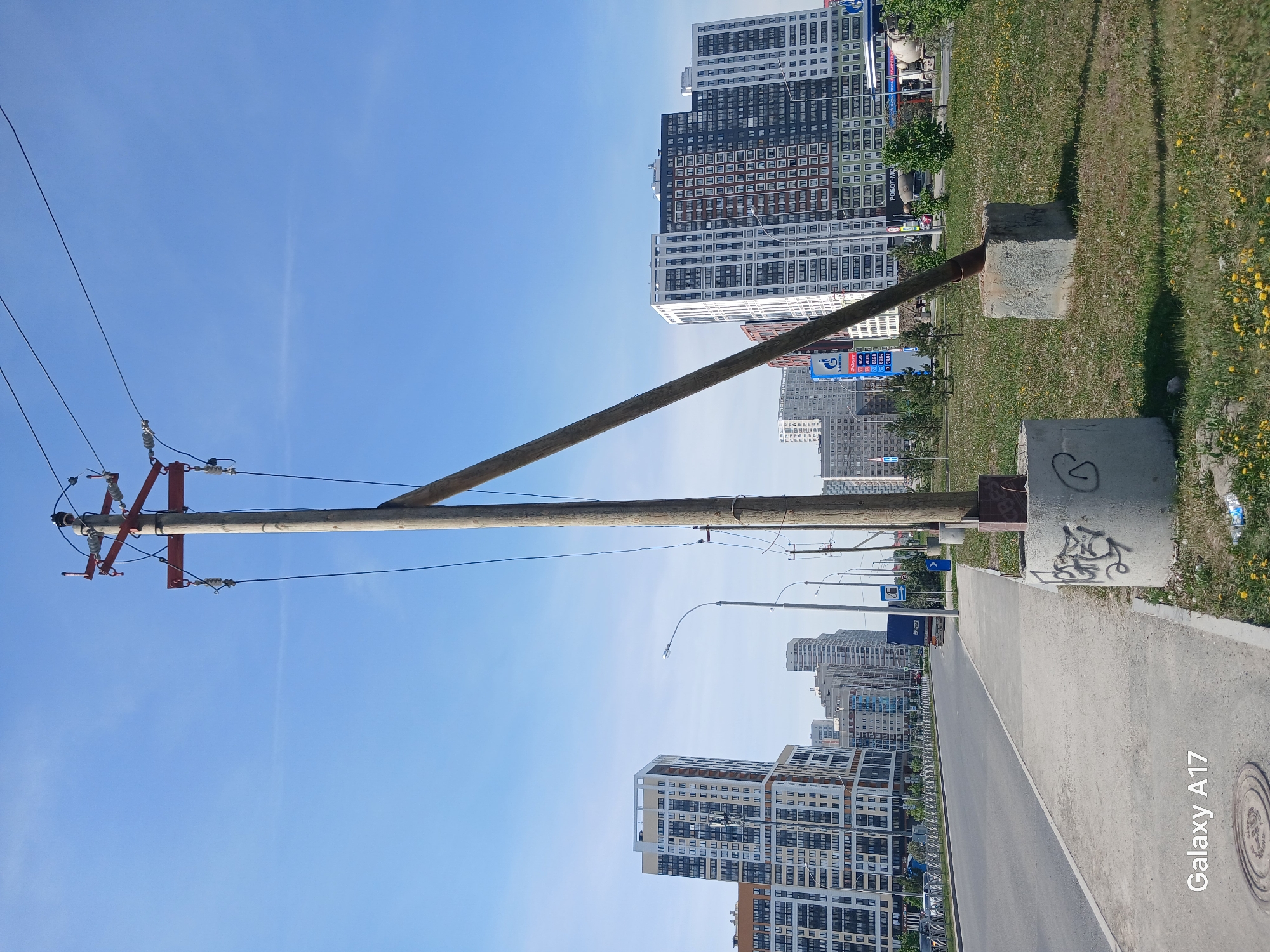 The image shows a street scene in what appears to be a modern urban area, likely Yekaterinburg, Russia. In the foreground is a utility pole with a diagonal support brace attached to concrete blocks at the ground level. The pole is equipped with electrical hardware and wires extending towards the upper left. The right side of the image features a sidewalk adjacent to a roadway. Modern high-rise buildings dominate the background, suggesting a densely populated urban setting. The sky is a clear blue, indicating good weather conditions. There are patches of grass and vegetation present along the sidewalk and on the opposite side of the street. Graffiti is visible on the concrete blocks near the base of the utility pole.