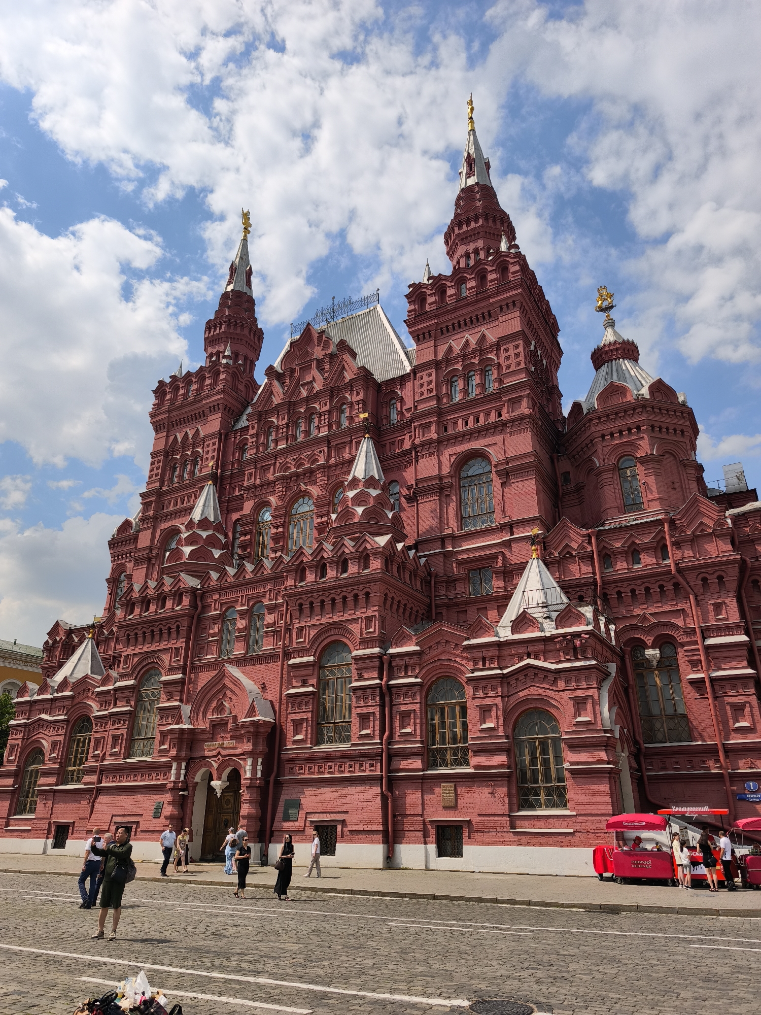 A wide-angle, eye-level exterior shot captures the State Historical Museum in Moscow, Russia, under a partially cloudy blue sky. The building, made of red brick, features ornate architecture with multiple towers, spires topped with gold ornaments, and arched windows. Several people are visible on the cobblestone street in front of the museum, some walking and others standing. Two red tourist kiosks with awnings are parked along the sidewalk. The overall lighting suggests a bright, daytime scene.