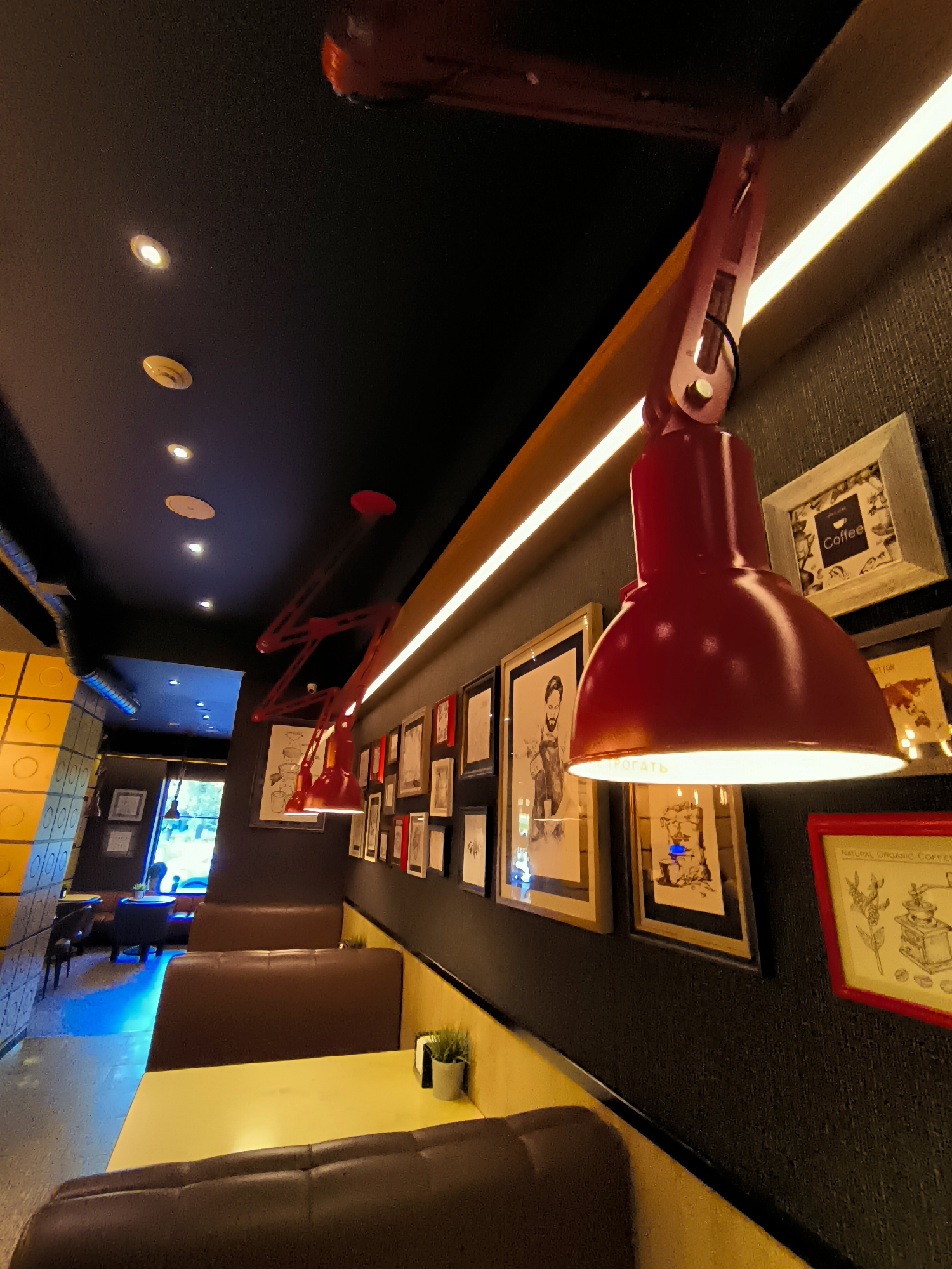 The image depicts the interior of a cafe, likely in the city of Karaganda, Kazakhstan, as indicated by the provided contextual information. The perspective is from a low angle, looking upwards towards the ceiling and along a wall adorned with framed pictures and red pendant lamps. The ceiling is black, with recessed lighting fixtures. A horizontal strip of warm-toned light runs along the upper part of the wall. The wall itself has a textured, dark finish.
The foreground features brown leather booth seating with yellow tabletops. Further into the cafe, more booths are visible, illuminated by a vibrant blue light. On the yellow wall, a small potted plant is placed near a napkin dispenser.
The wall display consists of an array of framed pictures. Many of these appear to be drawings or prints, some depicting figures or scenes, and one framed with text that includes "Coffee." Another framed picture on the right has a drawing of a coffee grinder and the text "NATURAL ORGANIC COFFEE."
Two red, articulated pendant lamps are prominent, hanging from the ceiling and directed downwards, providing illumination. One lamp is closer to the foreground and its shade is brightly lit from within. Another similar lamp is visible further down the wall.
The overall ambiance suggests a modern and somewhat artistic cafe setting. There are no people visible in the immediate foreground or middle ground, but a window or doorway at the far end of the cafe offers a glimpse of outdoor greenery and possibly some seating. The architectural elements include textured walls, booth seating, and overhead lighting, creating a cozy yet contemporary atmosphere.