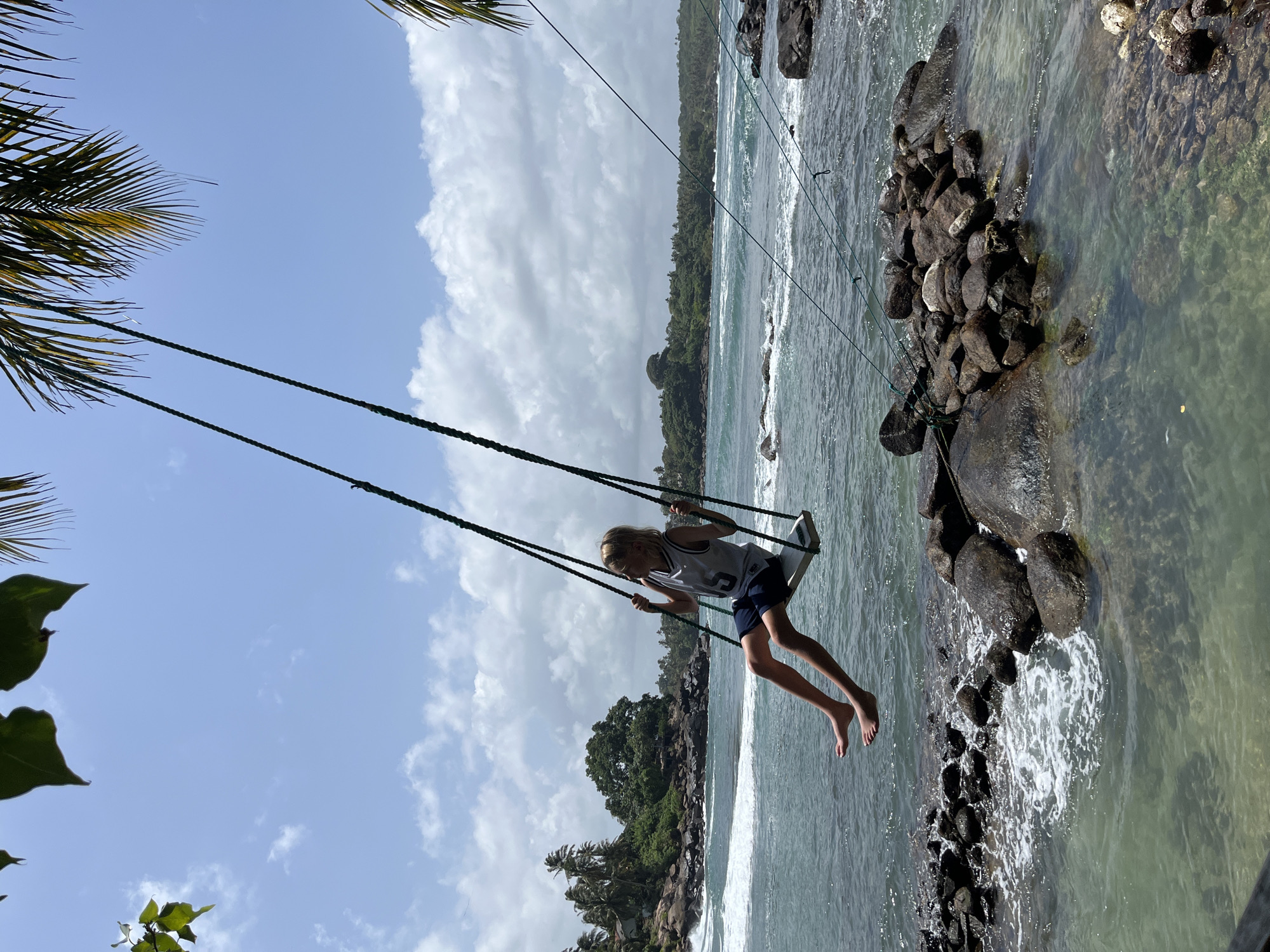 A young person with blonde hair is swinging on a rope swing over a rocky shoreline in Dondra, Sri Lanka. The person is wearing a grey tank top with the number "5" and dark blue shorts. The swing is suspended from palm trees and appears to be over the ocean. The water is a turquoise color, and waves are crashing against the rocks. In the background, there is a lush green coastline with palm trees and buildings. The sky is blue with scattered white clouds. The image captures a moment of joy and freedom as the person enjoys the swing in a tropical paradise.