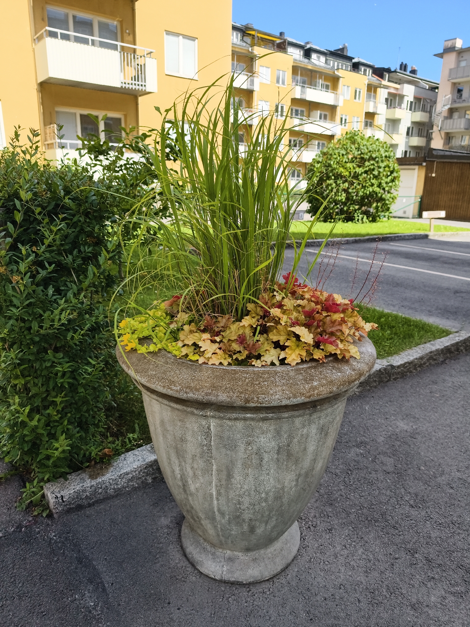The image depicts a large, aged concrete planter filled with a variety of plants. The planter is situated on a paved surface, possibly a sidewalk or parking lot, with a grassy area adjacent to it. In the background, there are several multi-story apartment buildings, painted in shades of yellow and white, with balconies and windows visible. A section of dense green foliage from a bush or hedge is seen to the left of the planter. The sky is clear and blue.
Based on the architectural style of the buildings and the overall setting, this scene is characteristic of residential areas found in Swedish cities, including Linköping. The apartment buildings suggest an urban or suburban residential environment. The presence of well-maintained planters and greenery is common in such locations.