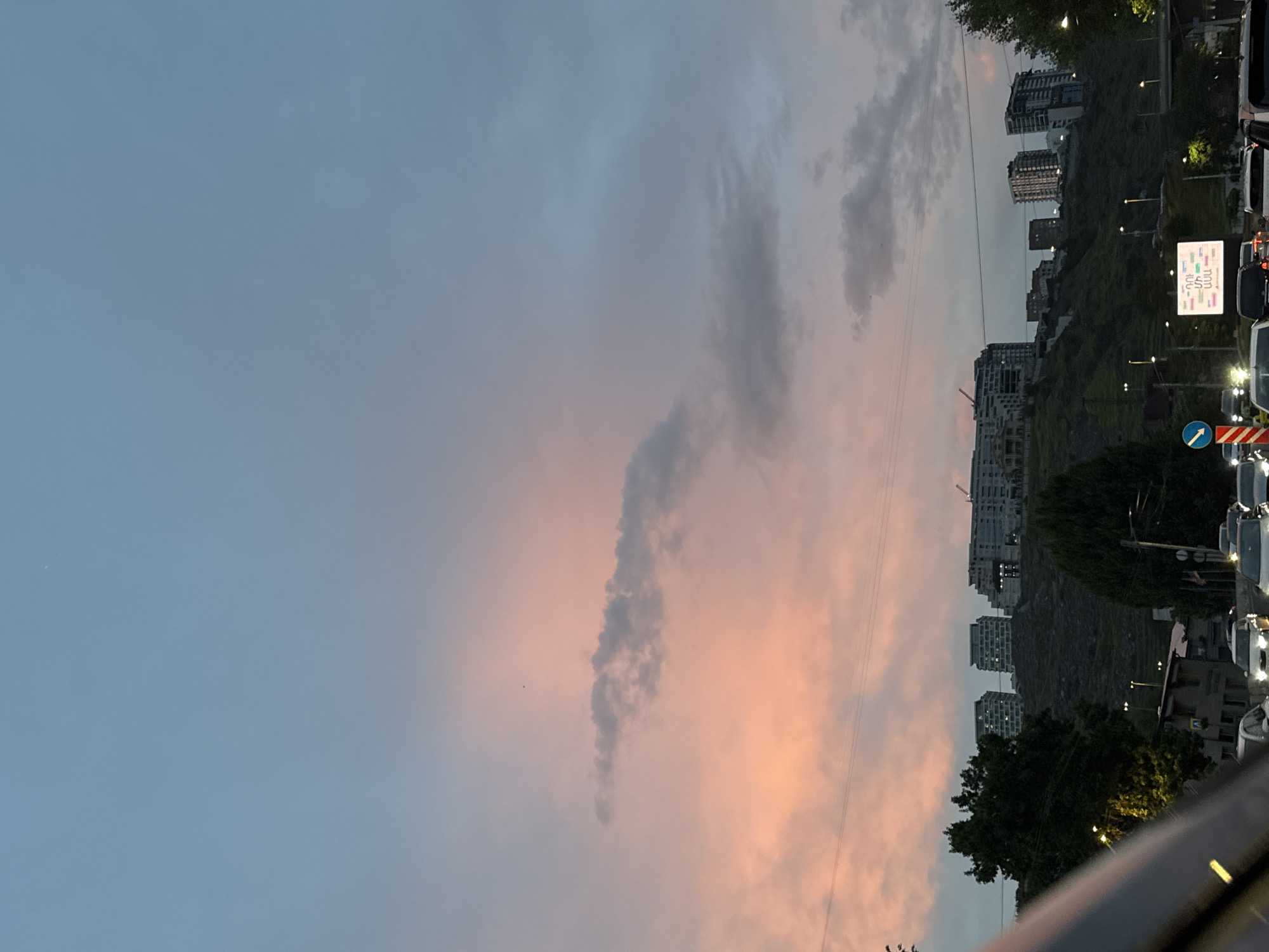 Captured at dusk in Yerevan, Armenia, the image presents an urban tableau against a sky streaked with pink and grey clouds. On the right, the cityscape features mid-rise buildings nestled on a hillside, street lamps illuminating the ascent, and lush trees dotting the landscape. A traffic jam is visible with an array of vehicles lining the roadside. Signage is present, but the text is illegible from this distance. The horizon is defined by the silhouettes of the buildings and vegetation against the fading light. The photograph is taken from a low vantage point, partially showing the hood of a vehicle in the foreground, suggesting the perspective of someone inside a car.
