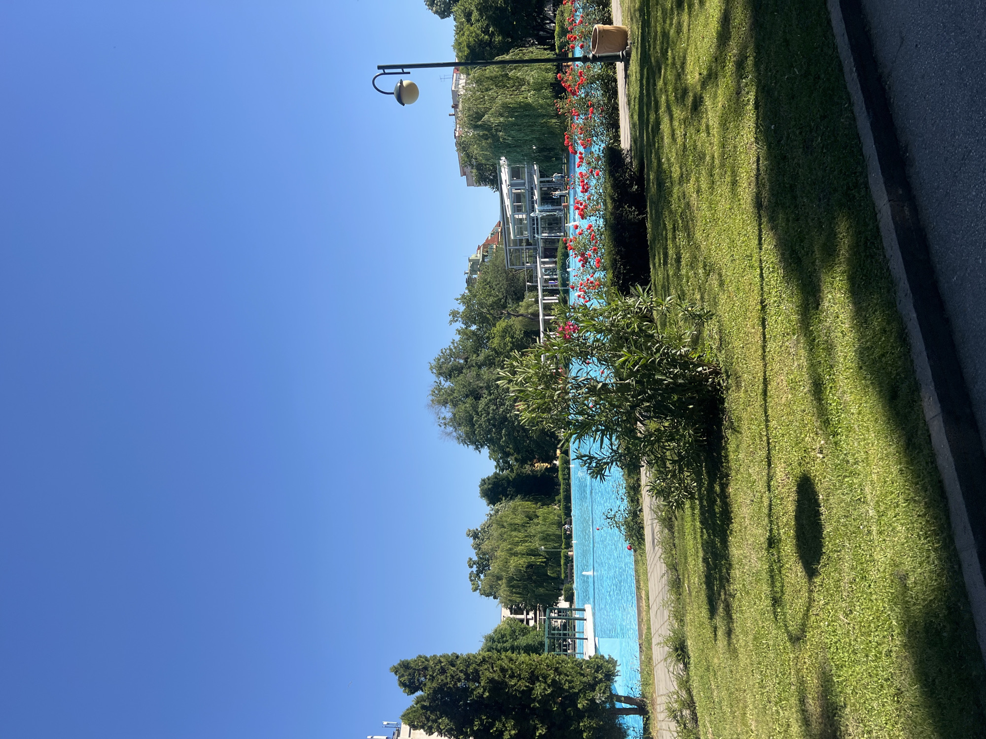 The image captures a sunny day at the Tsar Simeon Garden in Plovdiv, Bulgaria. A long, rectangular, bright blue pool stretches through the frame, bordered by lush green grass on one side and a walking path on the other. Red flowers and green shrubbery line the edge of the pool. The water is disturbed by the presence of multiple fountains. On the other side of the pool are trees, a white structure with metal beams, and buildings in the background. A street lamp with a spherical light fixture stands near the pool. The sky is a clear, unblemished blue.