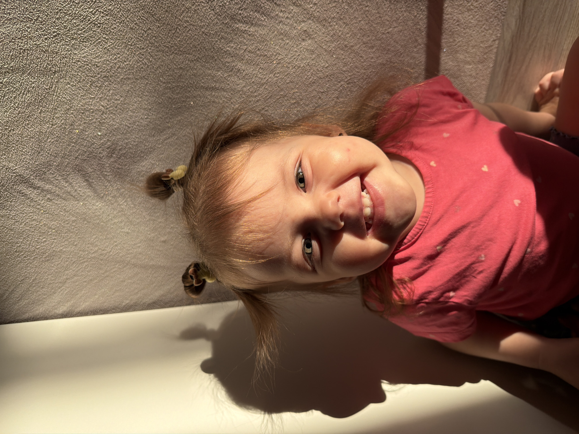 A young child with light skin, blonde hair styled in pigtails with brown hair ties, and wearing a pink shirt with small white hearts, is lying on a surface with a textured gray background. The child is smiling and looking directly at the viewer. The scene appears to be indoors, with sunlight casting shadows on the surface. The location is not discernible as Reshetnikovo, Russia, based on the visual information provided.