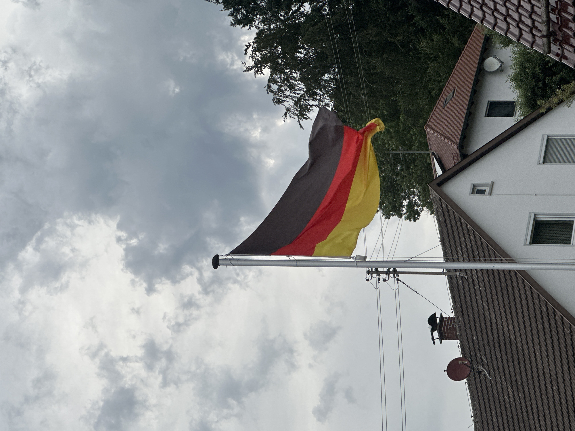 The image shows a German flag waving on a flagpole against a cloudy sky. In the foreground, the roofs of two houses are visible, one with a satellite dish and a chimney. Trees are seen in the background. There are no people or animals in the image. The scene appears to be a residential area.