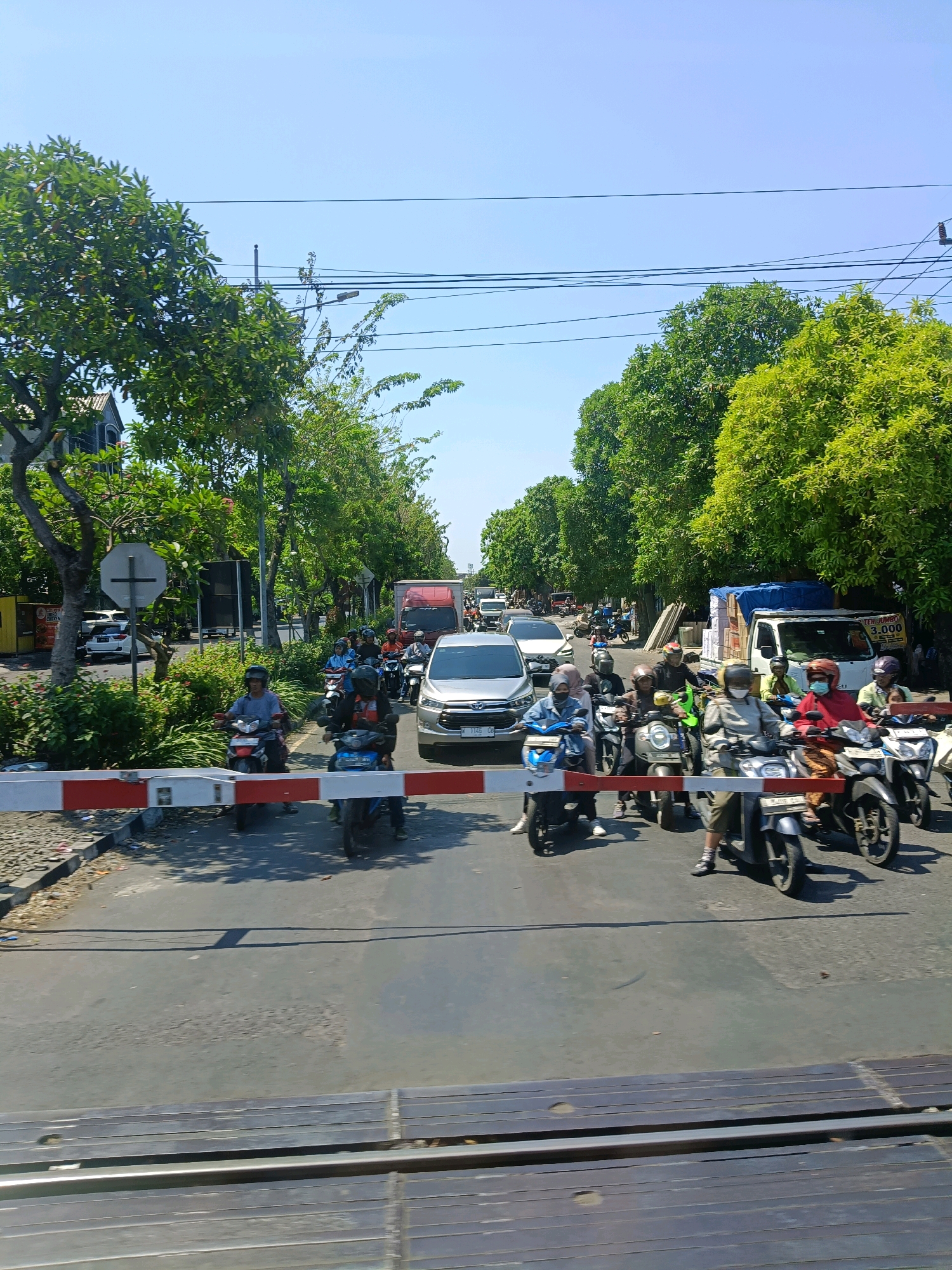 In Surabaya, Indonesia, on a sunny day, a traffic jam is occurring at a railway crossing. A red and white barrier is lowered across the road, halting traffic. Numerous motorcycles, most with riders wearing helmets, are stopped behind the barrier. Some motorcycles are moving forward, attempting to get through. A silver car and a red truck are also visible in the traffic. Lush green trees line both sides of the road, and power lines are strung across the bright blue sky. In the foreground, train tracks are visible, indicating the presence of the railway crossing. The scene depicts a typical urban traffic scenario in Surabaya, with the challenges of a busy railway crossing.