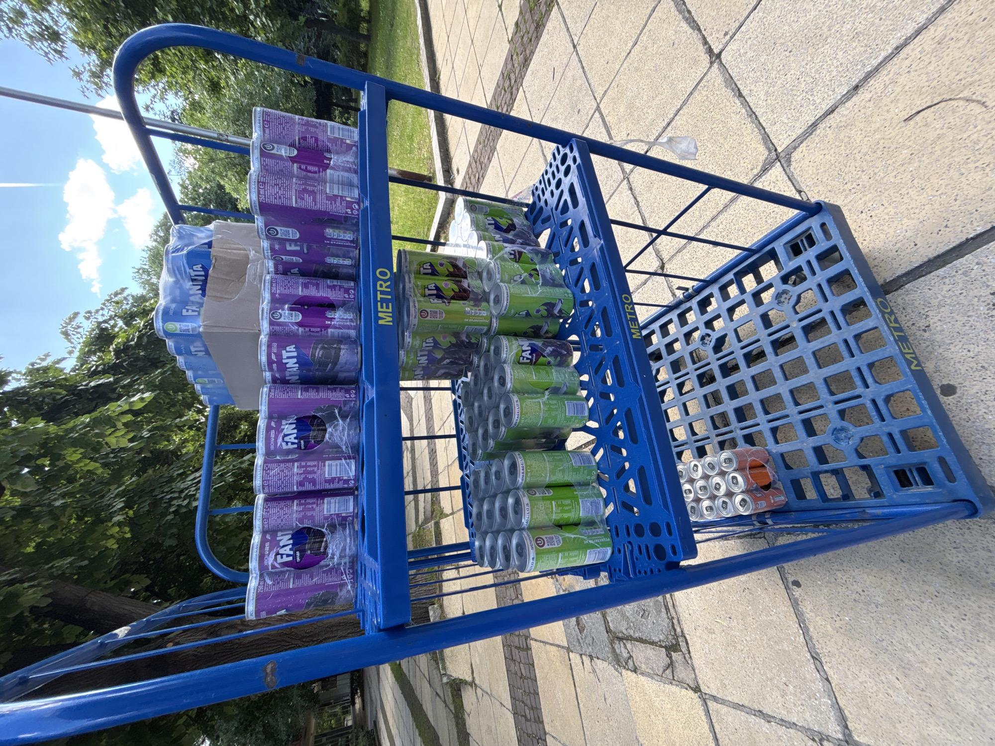 A blue metal and plastic Metro brand shopping cart stands outdoors on a paved area, likely a sidewalk, in Varna, Bulgaria. The cart is filled with stacks of canned beverages. On the left side of the cart are multiple packs of purple-labeled Fanta cans. Next to them are green-labeled Fanta cans. At the far right end of the cart is a stack of smaller, red-labeled cans. The setting appears to be near a park or green space, with trees visible in the background. The sky is bright, suggesting it is daytime. The overall impression is that the cart is loaded with various drinks, possibly for a store or event.