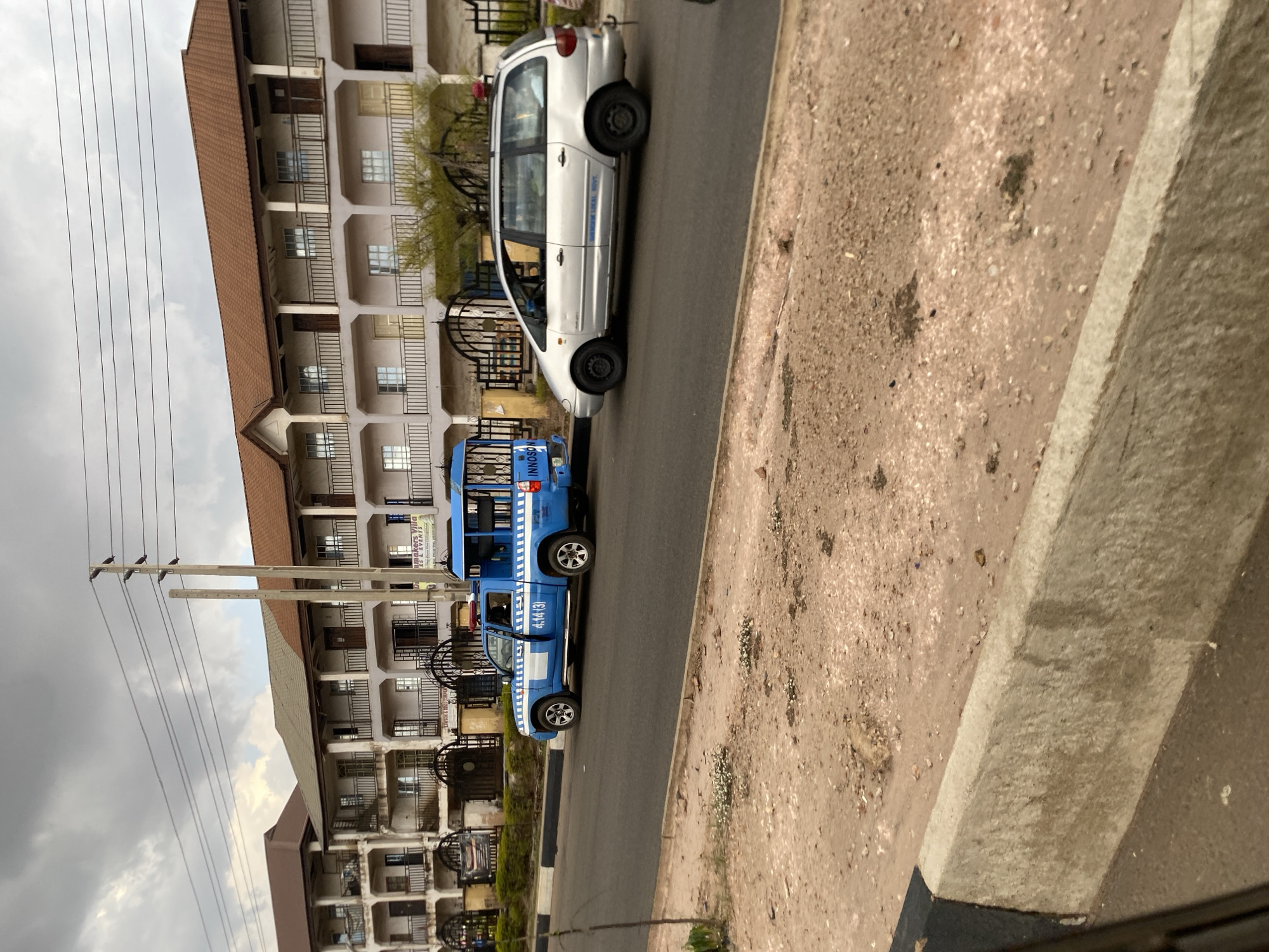 The scene depicts a street in Gura Top, Nigeria. A multi-story building with multiple balconies and windows stands to the left. Electrical poles with wires extend across the sky. Parked on the street are two vehicles: a silver minivan and a blue and white minibus. The foreground features a curb and a dirt shoulder.