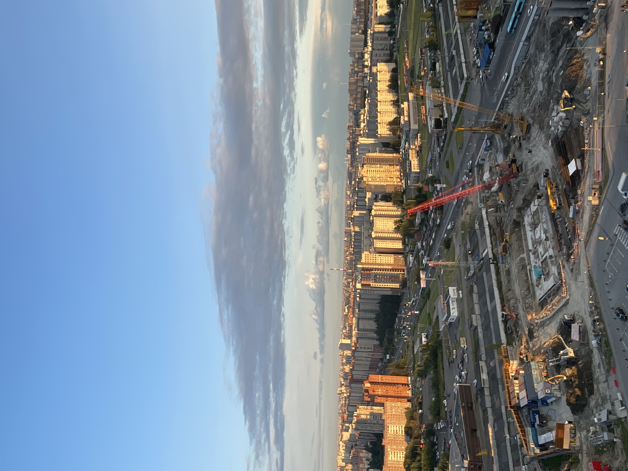This is an aerial shot of a city under construction. In the foreground, a construction site is actively being worked on, with cranes, excavators, and stacks of building materials visible. Several modular construction trailers are also present. Further in the background, a densely populated urban area with numerous apartment buildings stretches towards the horizon. A highway or major road with traffic is visible running through the city. The sky is a clear blue with scattered clouds, and the late afternoon or early evening sun casts a warm glow on the buildings.
