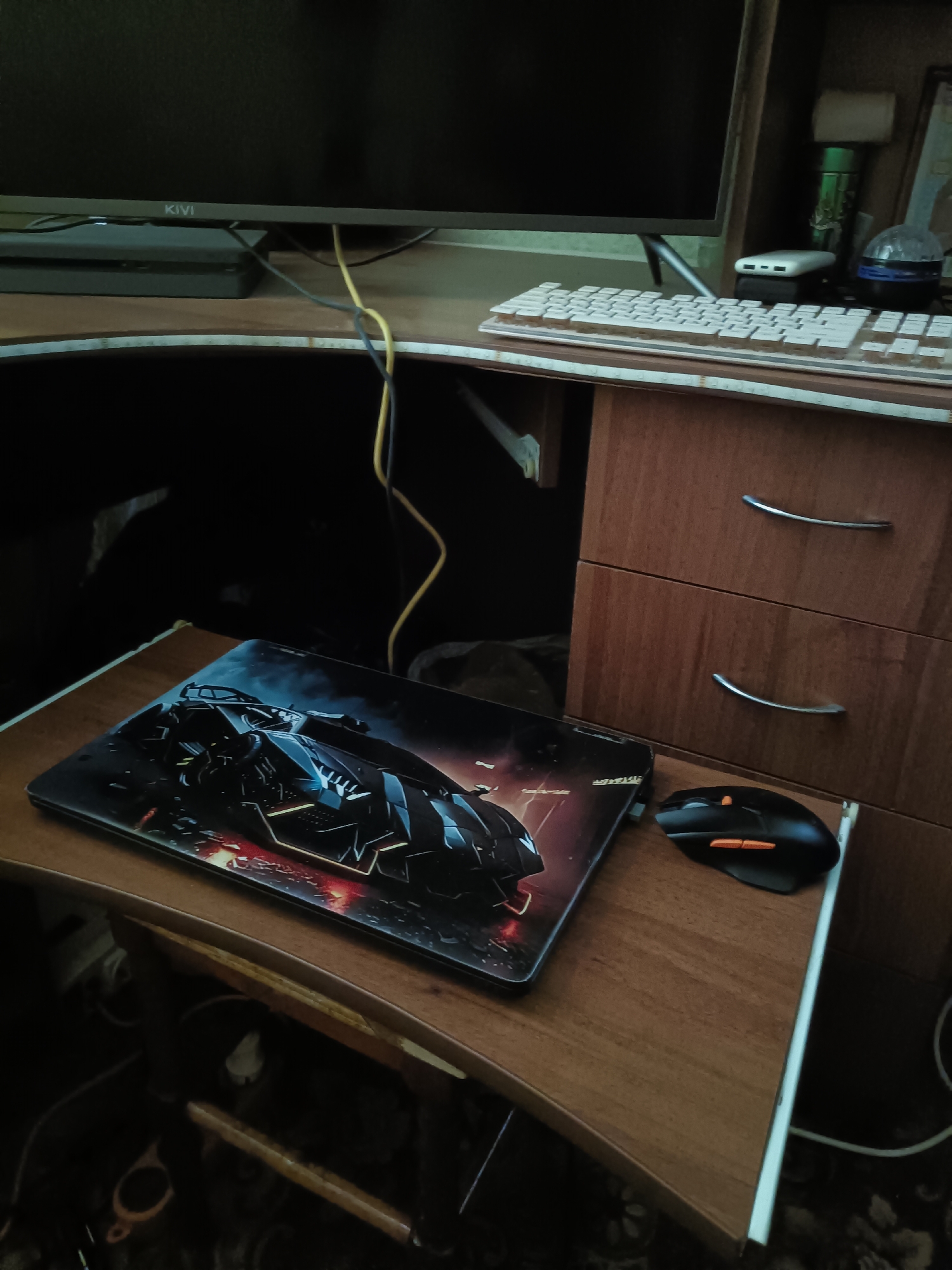 The media file displays a close-up, eye-level shot of a desk with a computer setup. The desk itself is made of wood and has a curved front edge. On the desk, there is a black computer mouse with orange accents, positioned to the right of a large gaming mousepad featuring a graphic of a dark, futuristic vehicle. A white keyboard is visible behind the mousepad, and a monitor is partially visible at the top of the frame. The monitor has a thin bezel and the brand name "KIVI" is visible on its lower edge.
Behind the monitor and keyboard, a wooden cabinet with two drawers is present. The drawers have silver, curved handles. A black cable and a yellow cable are seen running from under the desk and along the back of the setup. The scene appears to be indoors, likely in a home or office environment, given the presence of a computer and desk. The lighting suggests it could be daytime, though the interior setting makes it difficult to determine specific time of day or weather conditions. The overall impression is one of a functional workspace, possibly used for gaming or general computer use. No people are visible in the media file. The location context provided is Kotliareve, Ukraine.
