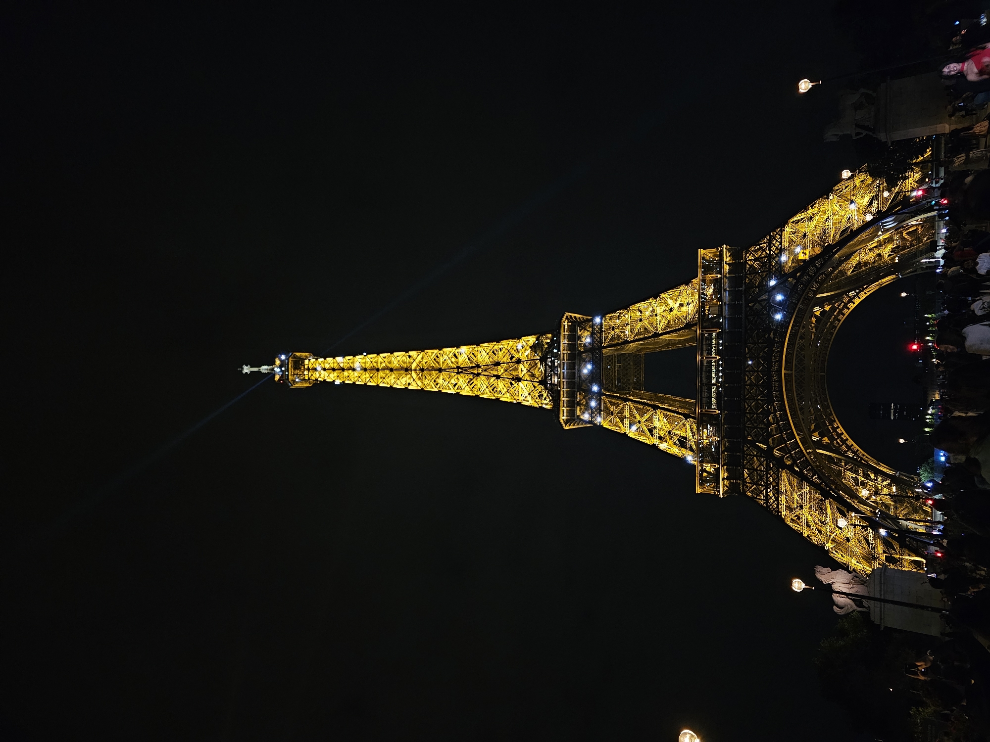 The image shows the Eiffel Tower in Paris, France, at night. The tower is illuminated with golden lights, and a bright light beam projects from the top. Crowds of people stand near the base of the tower. Streetlights and other architectural features are visible. The overall environment is dark, with the lit tower and surrounding lights creating a vibrant contrast.
