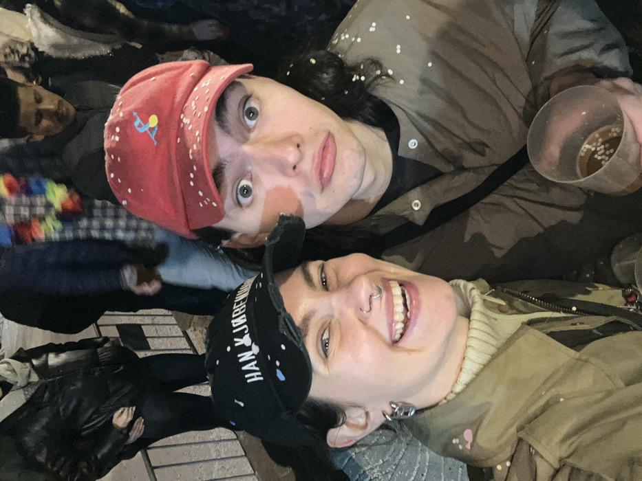Two people pose closely for an upward-angled selfie at night. The person on top wears a red cap with white specks and an illustration of a person skiing. They have wide eyes and a surprised expression. The person below wears a black cap with white and blue paint-like details and smiles widely, showing their teeth. They have a nose ring and metal ear gauges. Both individuals appear to be covered in confetti. A clear plastic cup with liquid and what seems to be confetti inside is visible in the top right. Other people are blurred in the background.