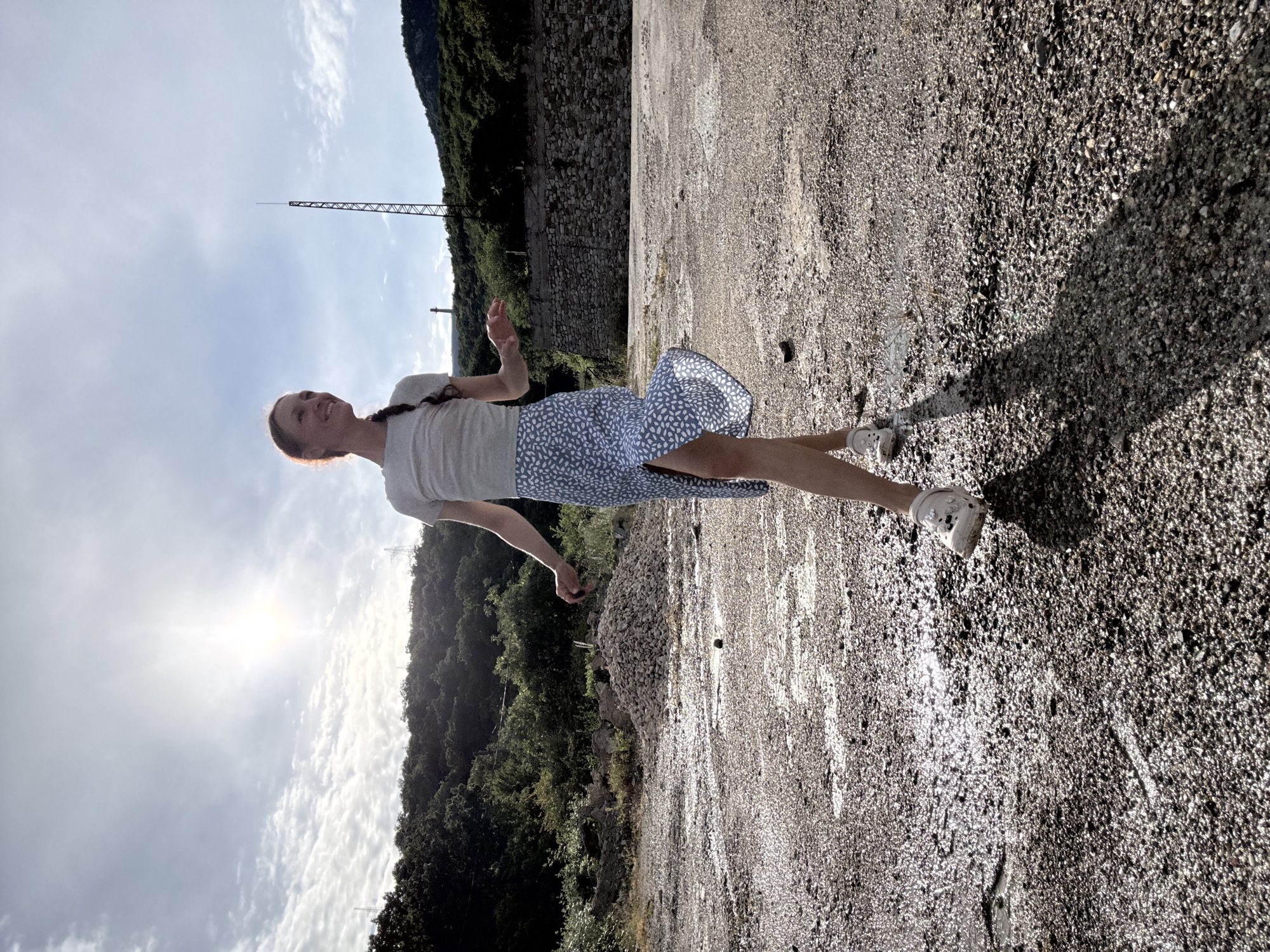 A young woman with long, braided red hair and fair skin is captured mid-stride in an outdoor, industrial-looking environment. She is wearing a light gray t-shirt and a blue and white floral print skirt that is split on the side, revealing her legs. Her feet are clad in white Croc-style shoes. The woman is looking to her left, with a slight smile on her face. Her right arm is extended outward, bent at the elbow, as if gesturing or in mid-dance.
The ground is a mixture of gravel and wet patches that reflect the sunlight, suggesting recent rain or a damp surface. In the background, to the left, a lush green hillside covered in trees rises, with sunlight glinting through the foliage. Further in the background, to the right, a tall, dark metal tower stands prominently against the sky, and beyond that, another smaller tower is visible. A stone wall, constructed from irregular blocks, runs horizontally across the middle ground, behind a pile of light-colored rocks. The sky is a mix of blue and white clouds, with the sun, partially obscured by clouds, shining brightly and casting a strong shadow of the woman on the ground. The overall scene appears to be located in an undeveloped or transitional area, possibly on the outskirts of a settlement.