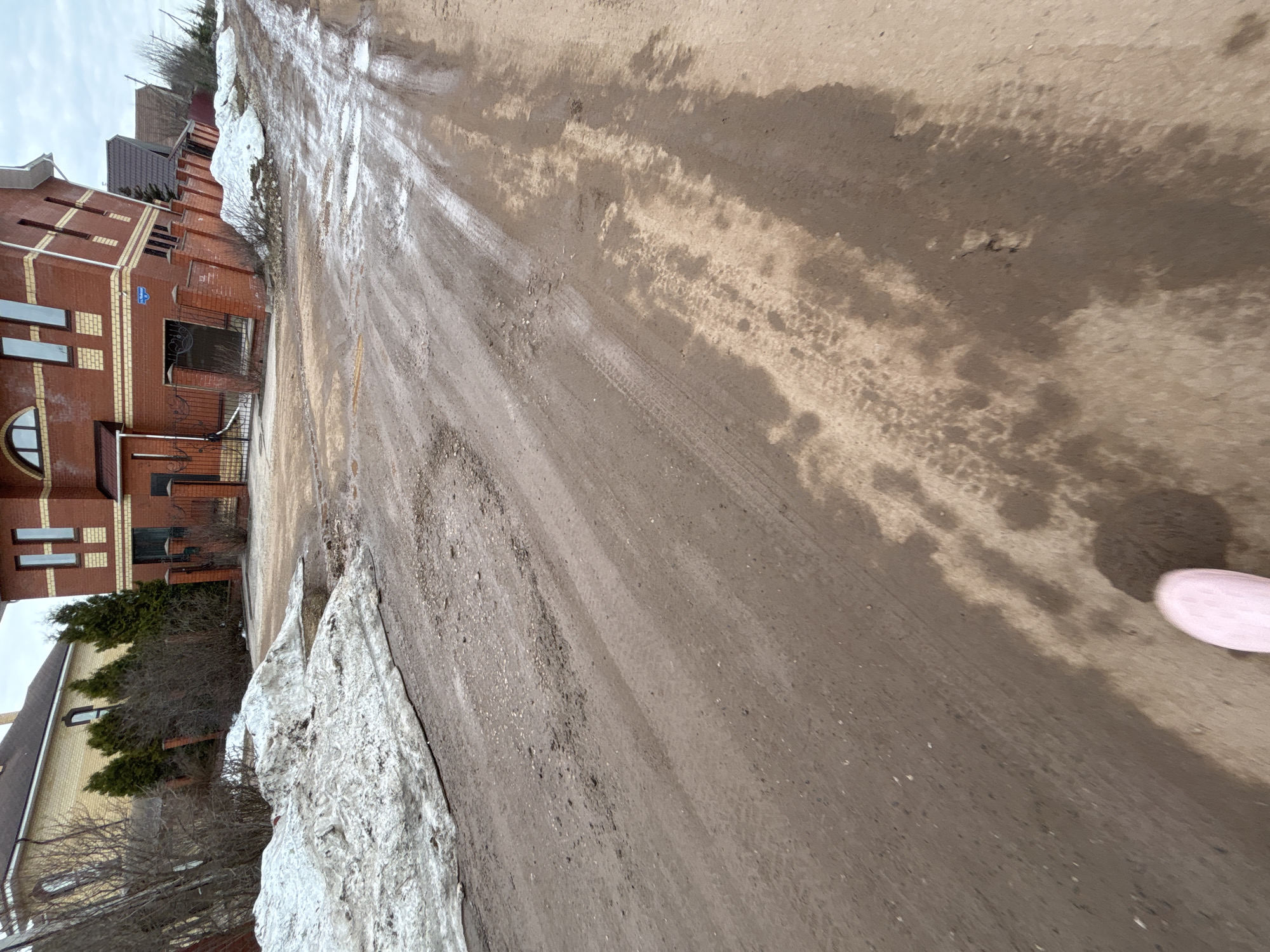 The image shows a muddy, rural street in Ферма, Russia, with melting snow along the roadside. A brick building is on the left. The street surface is uneven, with visible tire tracks. A glimpse of a pink object is in the lower right corner.