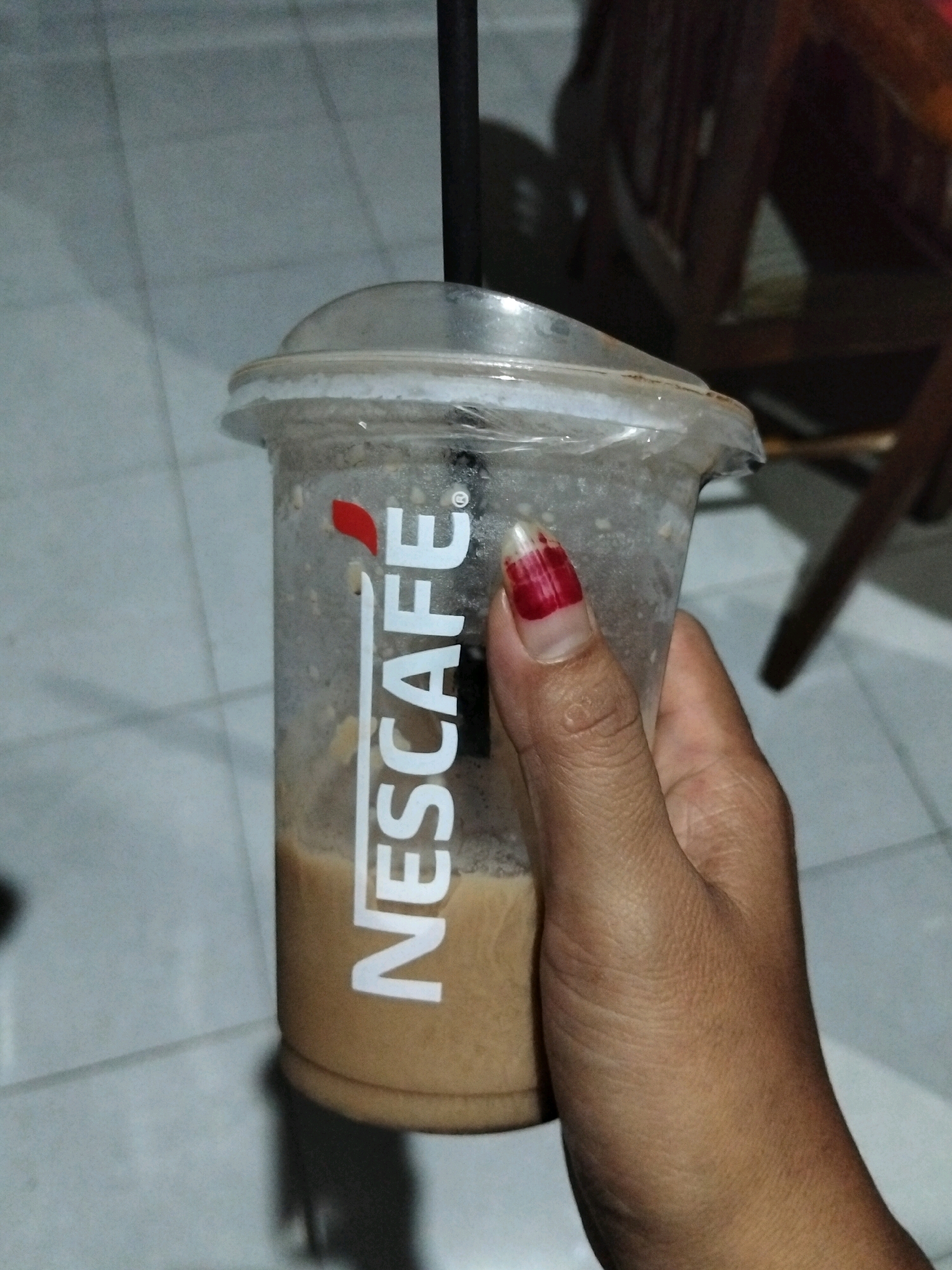 An outdoor, close-up, eye-level shot shows a person's hand holding a clear plastic cup with a Nescafé logo. The cup contains a light brown, frothy liquid. The hand has dark skin and painted fingernails with a red and white design. A black straw is visible in the cup. The background is blurred, showing a tiled floor and a wooden chair. The scene is located in Panimbang, Indonesia.