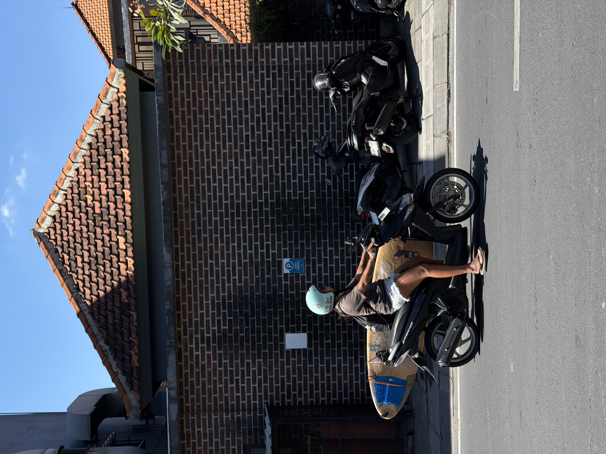 In North Kuta, Indonesia, a person wearing a light blue helmet and casual clothing is seated on a black scooter positioned on the side of a paved road. A surfboard is mounted on the scooter. Alongside the scooter, several parked motorcycles line the street. The backdrop consists of a brick wall and a building with a red-tiled roof. A "guest parking" sign is visible on the brick wall. The environment suggests a sunny day.