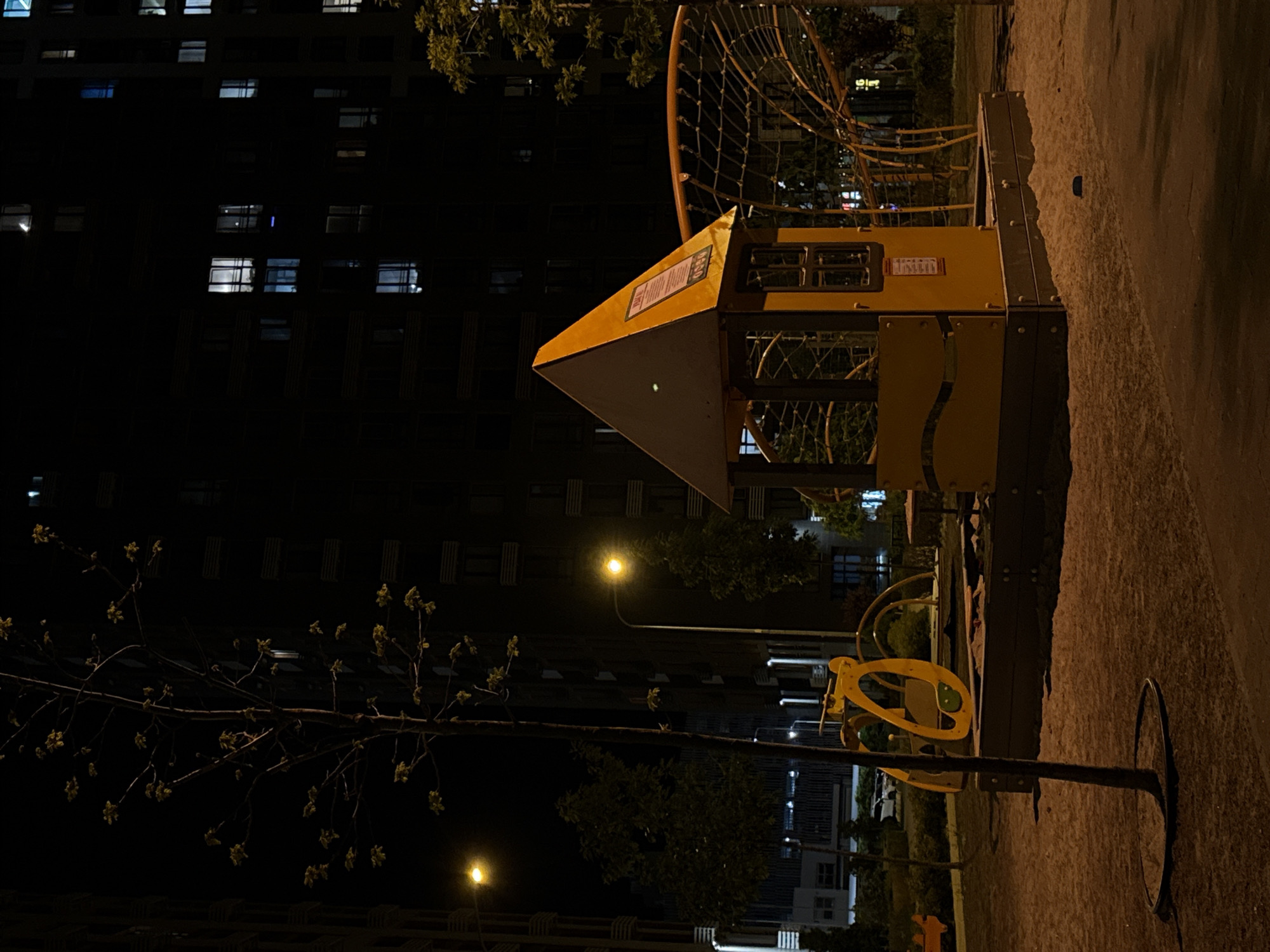 This nighttime image depicts a quiet residential area in Kyiv, Ukraine, showcasing a playground under artificial lighting. The scene is dominated by a dark, multi-story apartment building on the left, with numerous windows illuminated, indicating occupancy.
In the foreground and center, a children's playground features various equipment. A prominent yellow and brown structure, resembling a playhouse or climbing frame with a pointed, pyramidal roof, stands out. On its side, a rectangular sign with unreadable text is visible, likely providing informational details. To the right of this structure, a large, curved climbing net made of dark material extends upwards. Closer to the foreground on the right, another yellow playground element, possibly a small ride-on or spring rider, is present on the ground, alongside a tall, slender metal pole with a circular base.
The ground around the playground appears to be a mix of concrete or asphalt. Several streetlights emit a warm yellow glow, illuminating sections of the playground and surrounding areas, casting soft shadows. A young tree with small leaves or buds extends into the frame from the lower left, its branches silhouetted against the dark buildings. Other trees and bushes are scattered throughout the background.
The time of day is clearly night, with the sky completely dark. The weather appears clear and calm, with no visible precipitation. No people or specific activities are observed, giving the scene a tranquil and empty atmosphere.