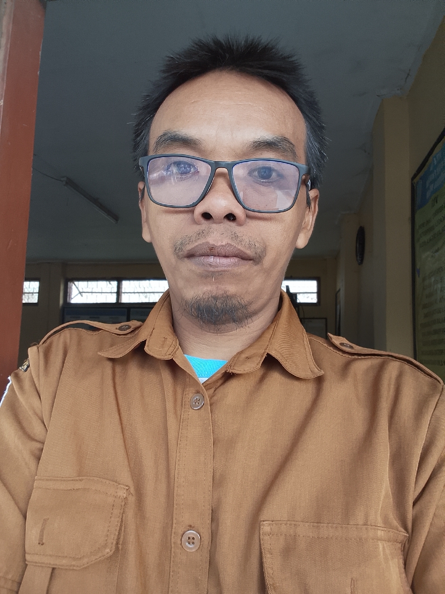 A medium shot portrait photograph shows a man wearing glasses and a brown collared shirt. The man is looking directly at the camera. His hair is short and dark. He has a goatee and mustache. The background appears to be indoors, with a window and a wall visible. The image is likely taken in Kepuh, Indonesia.