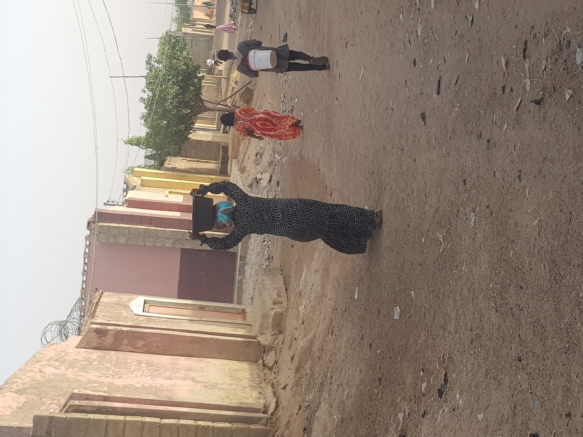 The image captures a dusty street scene in Dutsinma B, Nigeria. A woman wearing a long, black and white patterned dress and a blue headscarf is carrying a square, dark-colored object on her head. Further up the street, a woman in a red and orange patterned garment walks ahead of a person carrying a white cylindrical object on their shoulder. Buildings of varying heights and colors, primarily in earth tones, line the left side of the street. The street is unpaved and covered with dirt and debris. Overhead electrical wires stretch across the sky. The light is bright, suggesting a daytime scene.