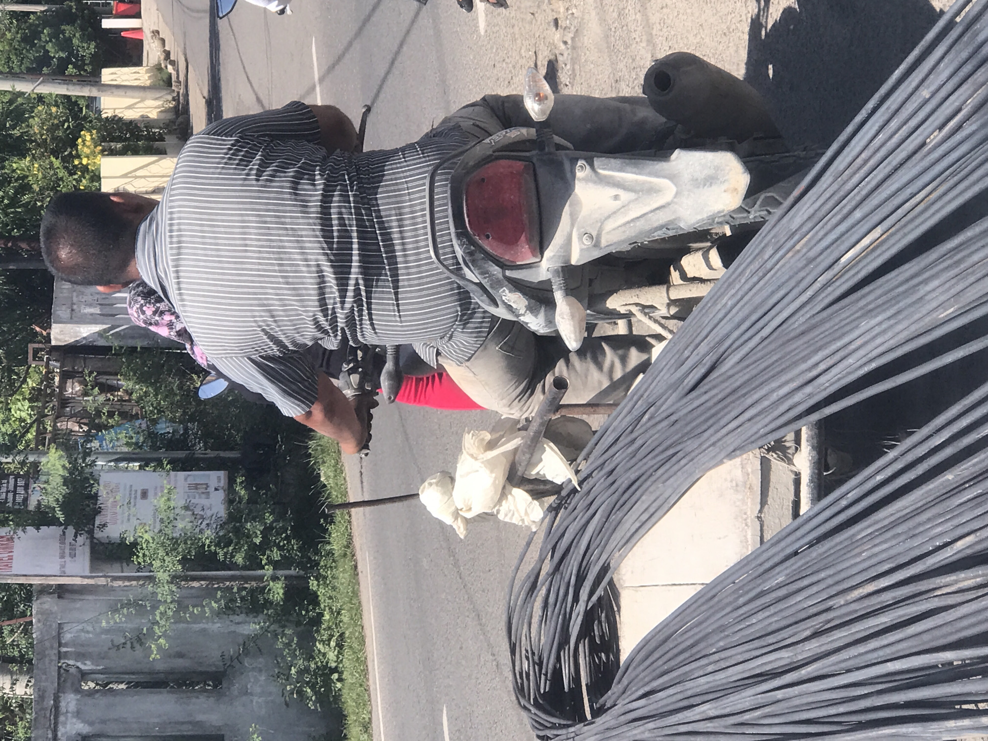 The image shows a man riding a motorcycle with a large bundle of reinforcing steel bars attached. The man is wearing a striped shirt and trousers and is facing the camera. The motorcycle appears to be heavily laden with the rebar. They are on a road. The city of Lhokseumawe, Indonesia is visible in the background. The background includes trees, buildings, signs, and other elements typical of an urban environment in Indonesia.