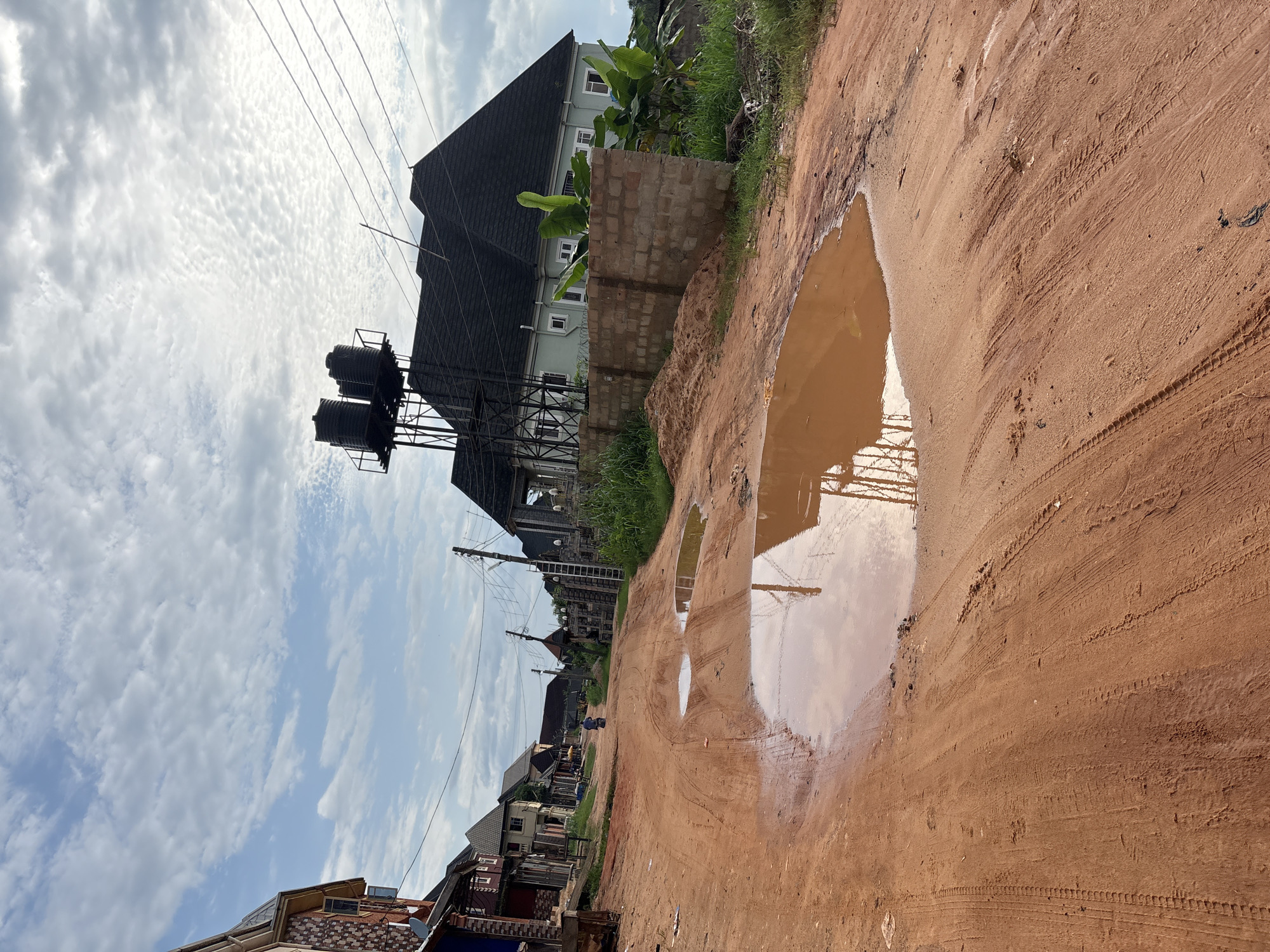 The image depicts a dusty, unpaved road in Obosi, Nigeria, with large puddles reflecting the sky and surrounding structures. On the right side of the road, a light green house with a dark roof is visible, with a brick wall in front of it. Behind the house, there are some tall green plants. Above the house, a metal structure supports three large black water tanks. To the left of the road, several multi-story buildings with varying architectural styles line the street. The sky is partly cloudy, with streaks of white clouds against a pale blue background. The road itself is textured with tire tracks from vehicles. In the distance, a single person is riding a motorcycle down the road. The overall scene conveys a sense of a developing urban or semi-urban area with infrastructure in progress, characterized by its earthy tones and the presence of water after rain.