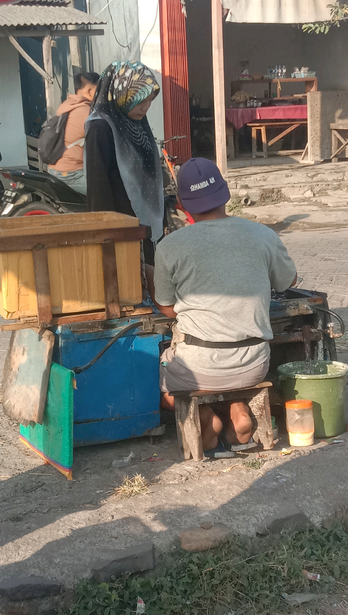 The image depicts a street scene in Curah Dukuh, Indonesia, focusing on a man sitting on a wooden stool behind a blue cart with a wooden container on top. He is wearing a grey t-shirt, grey shorts, and a purple cap with white lettering. His feet are bare. A woman wearing a colorful hijab and a dark garment stands to the left of the scene, looking down. Behind her, a person with a backpack is partially visible, standing near a motorcycle. The environment appears to be outdoors, with a building with red doors in the background. Various objects are present, including buckets, containers, and what appears to be a food cart setup. The ground is uneven and covered with dirt and some vegetation.