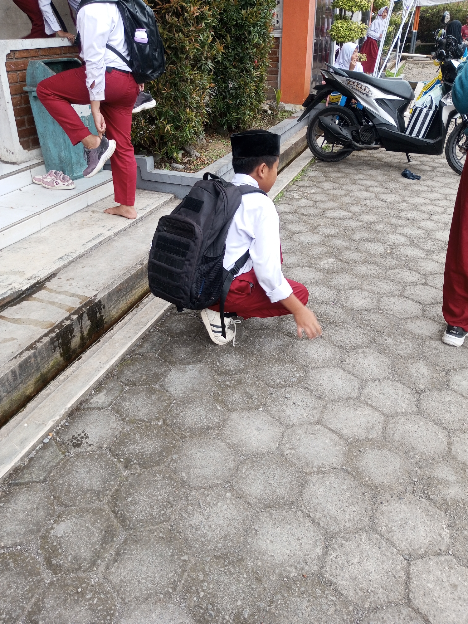 In the city of Kalibeber, Indonesia, a young student wearing a white shirt, black peci hat, and red pants is crouched on a honeycomb-patterned paved area. He has a black backpack on his back and is in the process of putting on a black and white sneaker. To his left, a pink sneaker lies on the ground near a green trash can. In the background, other students are visible, and a motorcycle is parked nearby. The scene appears to be an outdoor school environment with some vegetation.